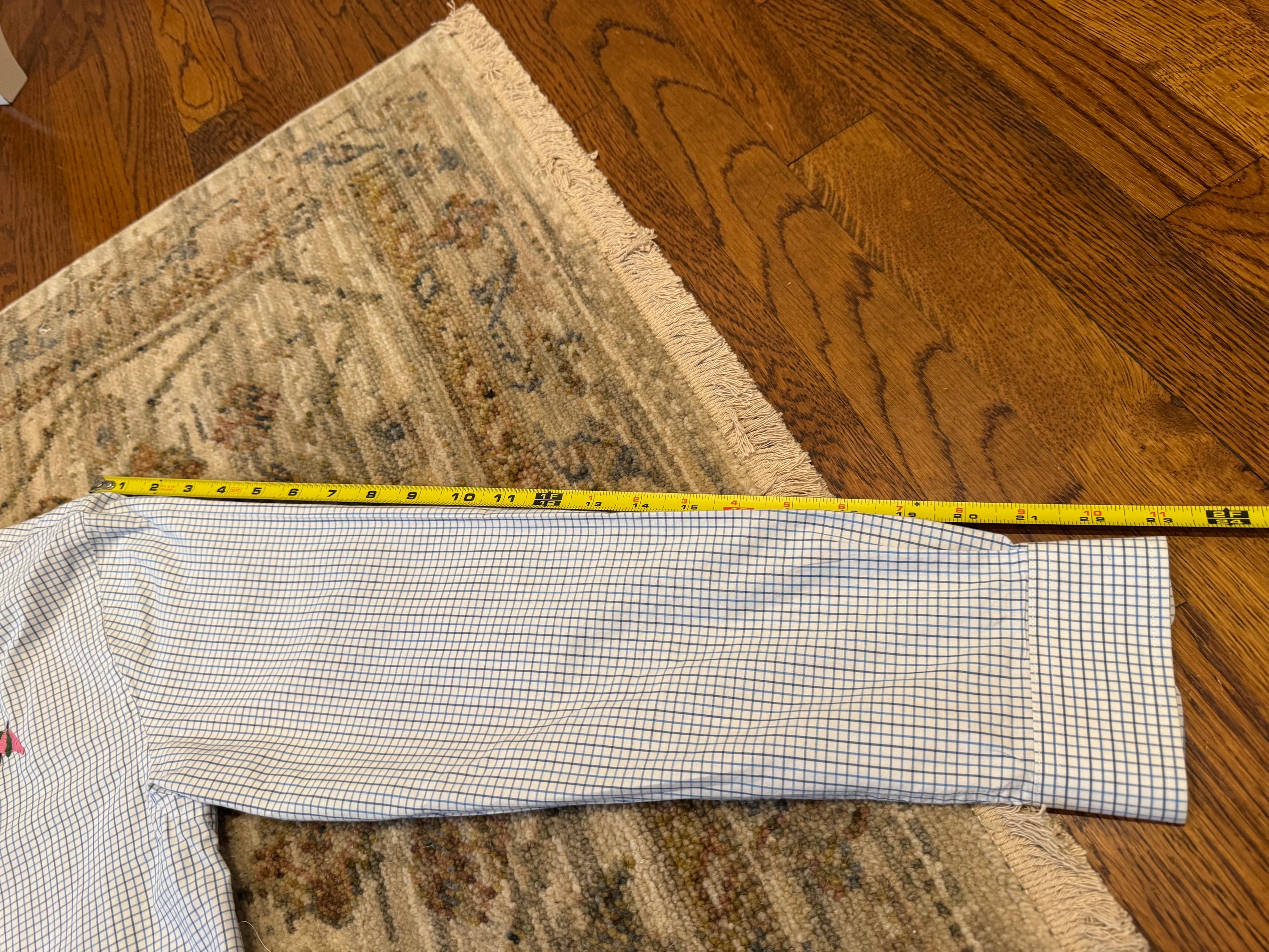 A long-sleeve button-up shirt laid flat on a patterned rug over a hardwood floor, with a yellow tape measure stretched across the sleeve to show its length.