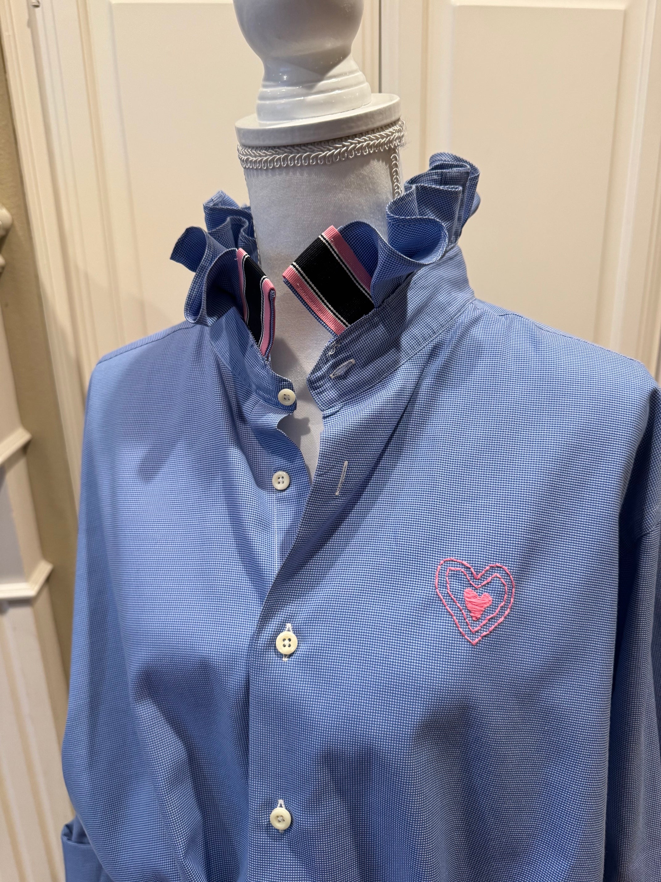 A light blue long-sleeve button-up shirt with a ruffled collar, displayed on a mannequin. The shirt features a pink embroidered heart on the left chest containing a small bird, and a striped necktie collar insert.
