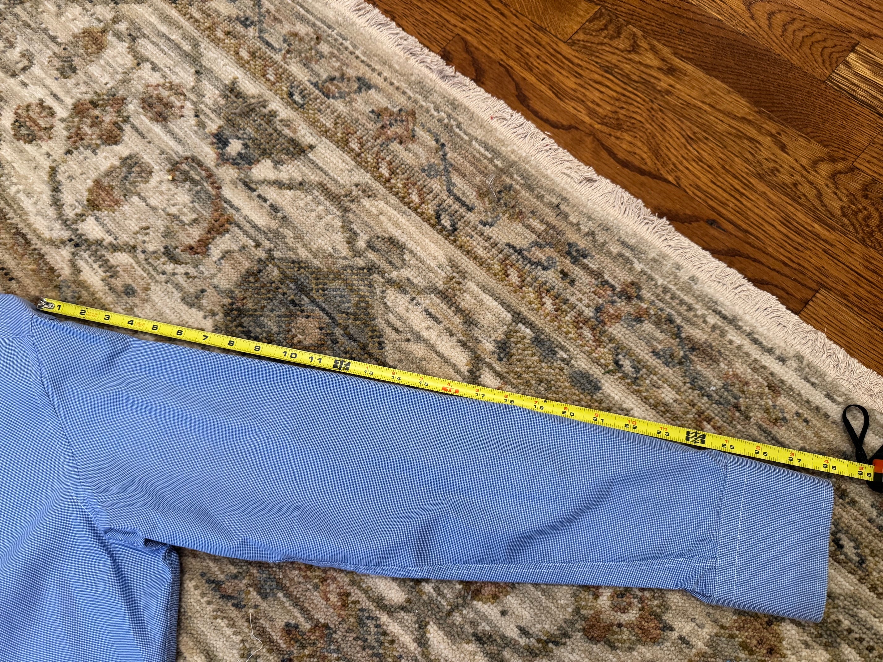 A light blue garment laid flat on a patterned area rug with hardwood flooring visible, and a yellow tape measure stretched across it to show dimensions.