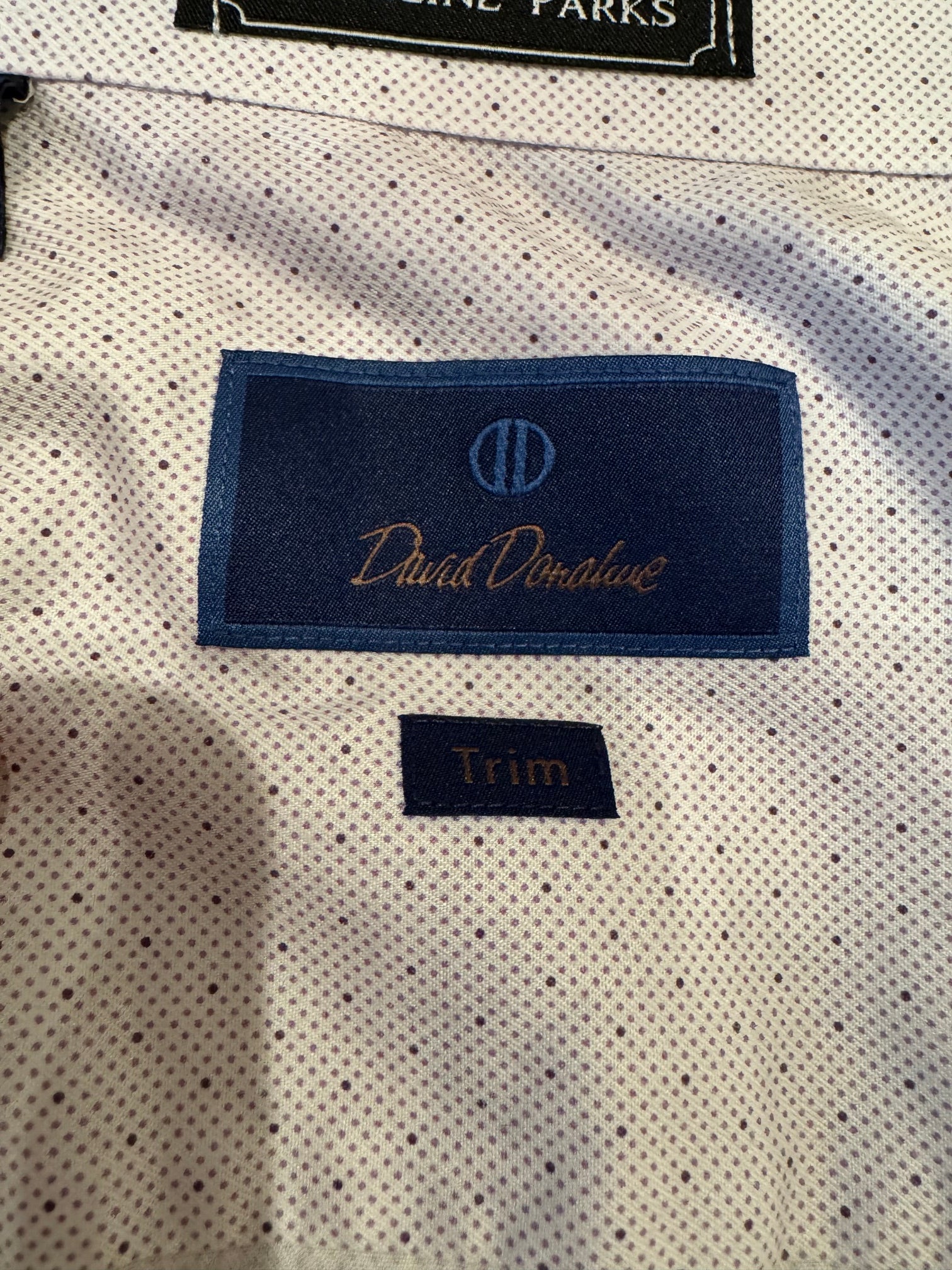Close-up of a white or off-white microfiber fabric with a fine black dotted pattern, featuring a blue embroidered brand label for 'David D'Evans' and a separate 'Trim' label.