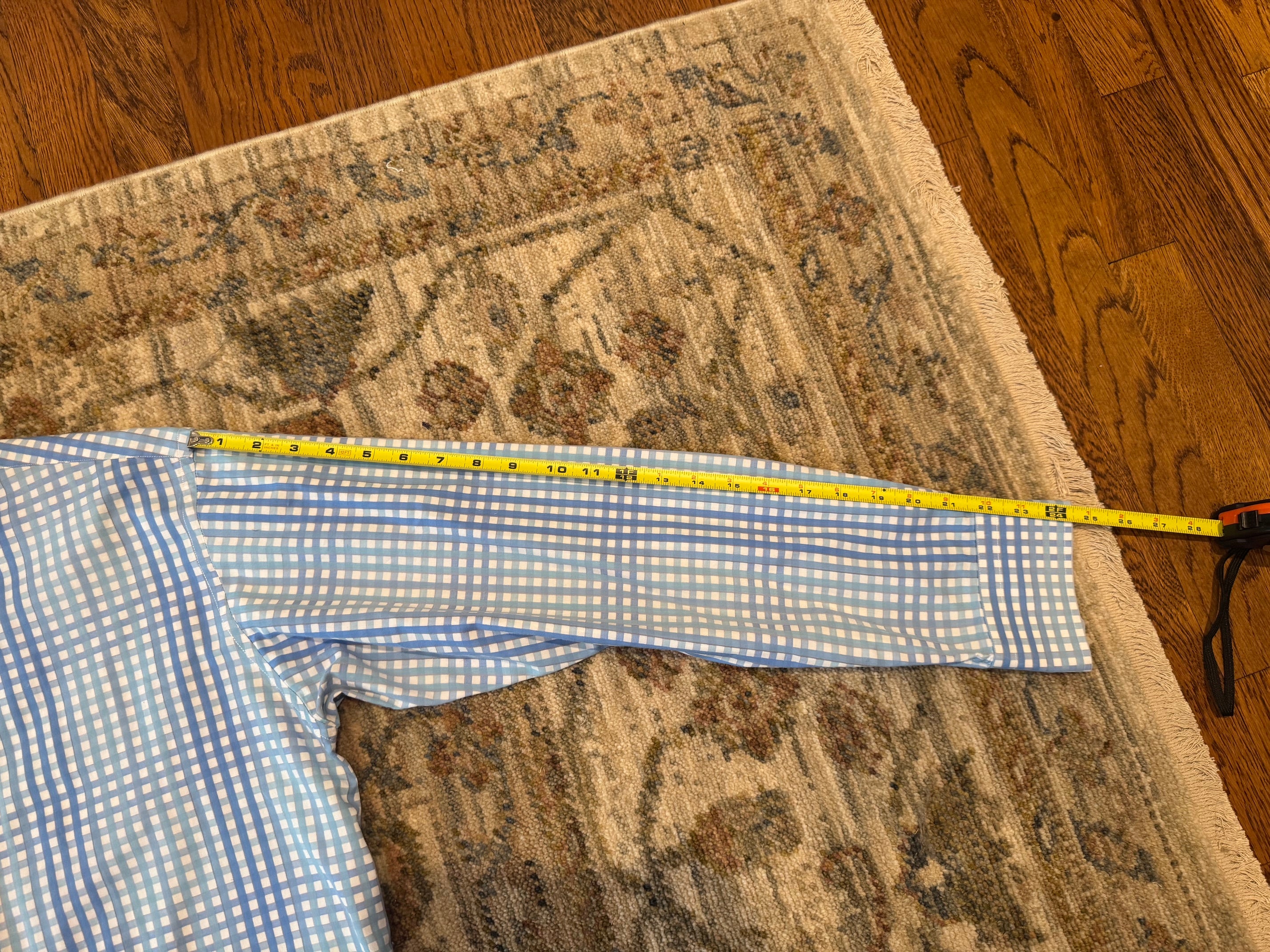 A blue and white gingham shirt laid flat on a patterned area rug with a yellow tape measure stretching across the sleeve to show its length, set on a wooden floor.