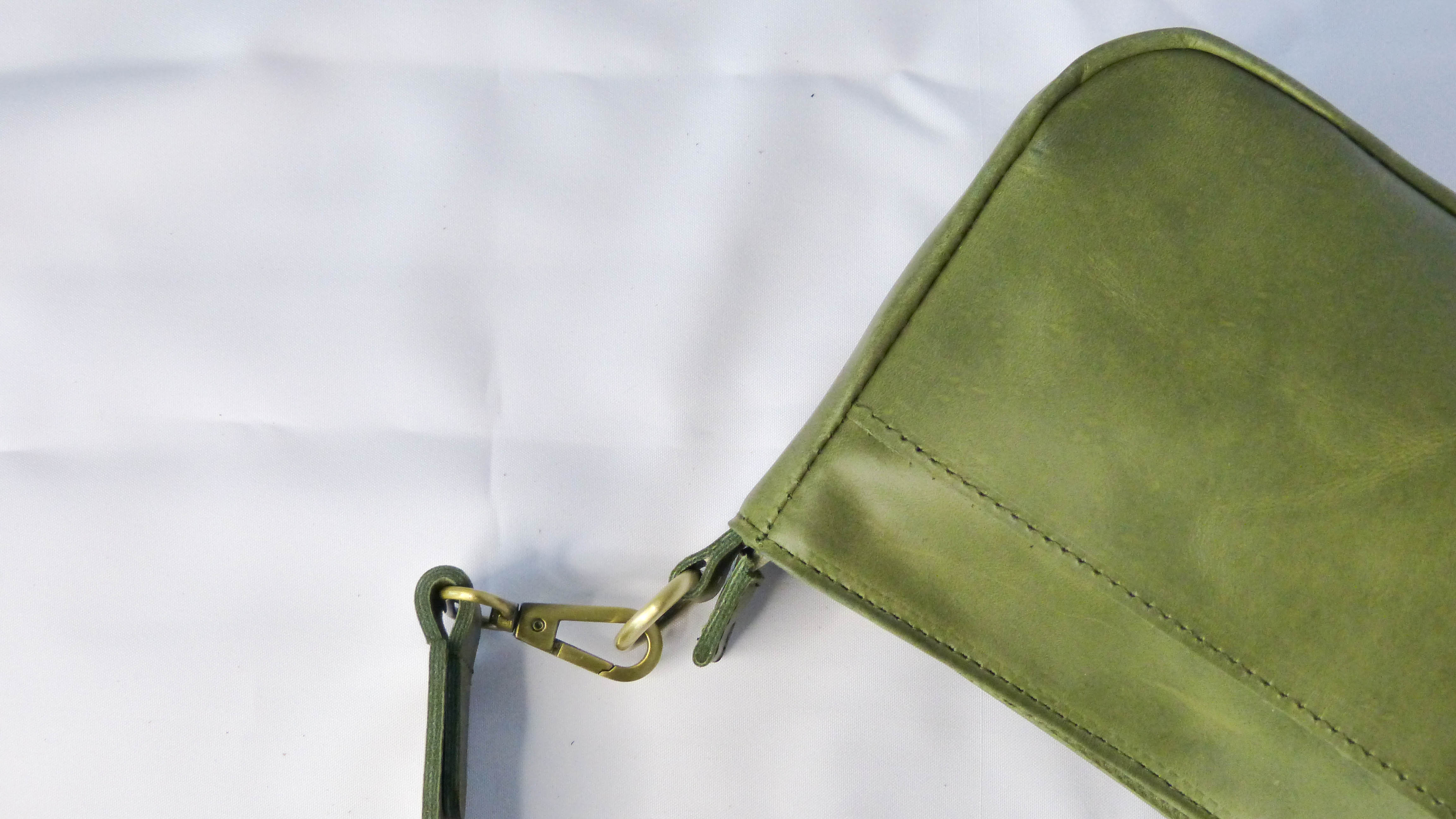A close-up of the Jessica Leather Purse in Olive, showcasing a green leather surface with a snap hook and metal clasp. Handmade by Ethiopian artisans, featuring a removable shoulder strap and zippered compartments.