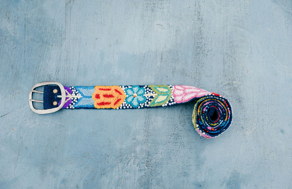 Navy Floral Embroidered Belt with bright florals, hand-loomed and embroidered by Peruvian artisans, displayed on a blue surface, showcasing detailed craftsmanship.