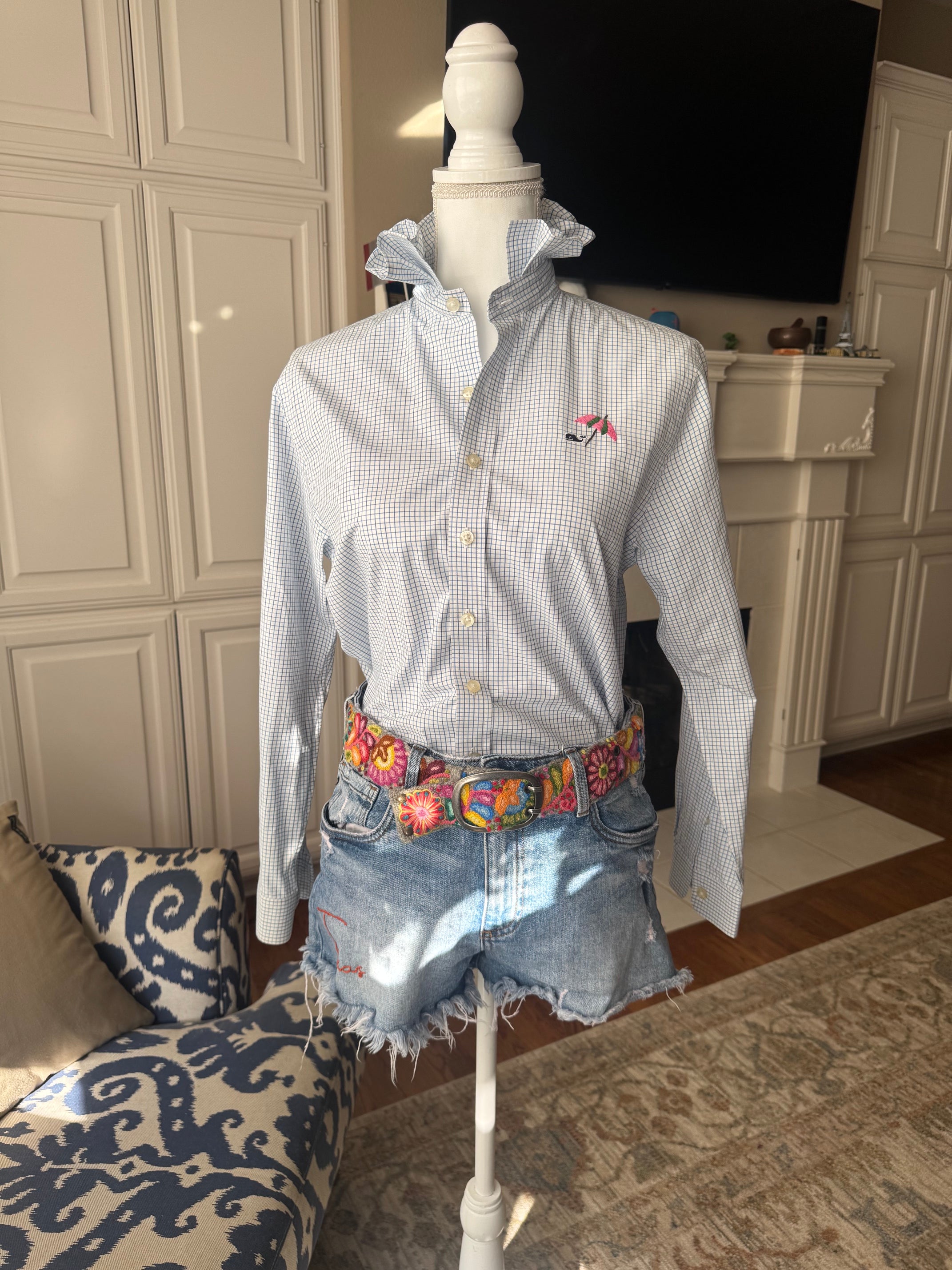 A light blue and white gingham long-sleeve button-up shirt with a small embroidered pink and green umbrella on the chest, styled on a white mannequin torso with denim shorts and a colorful patterned belt in a home interior setting.