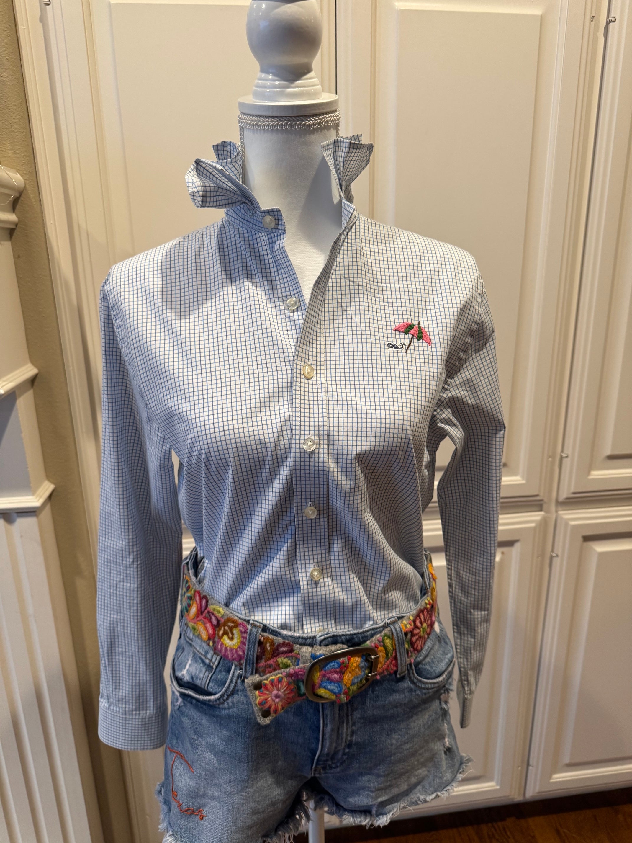 A light blue and white gingham long-sleeve button-up shirt with a small embroidered logo on the chest, styled on a mannequin with denim shorts and a colorful embroidered belt, set against a background of cream-colored wooden cabinets.
