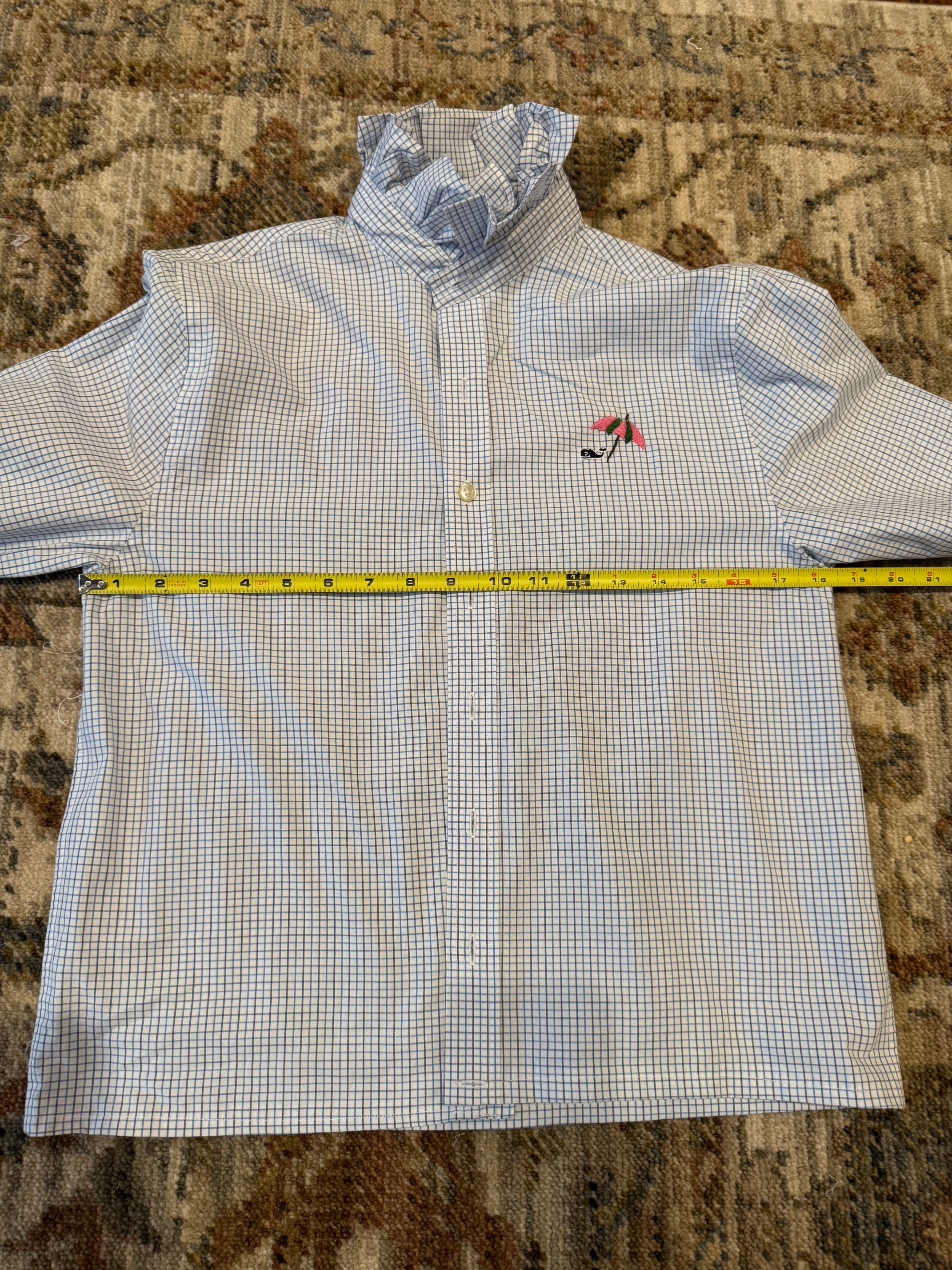 A light blue and white gingham long-sleeve button-up shirt laid flat on a patterned carpet, with a yellow tape measure stretched horizontally across the chest to show the size.