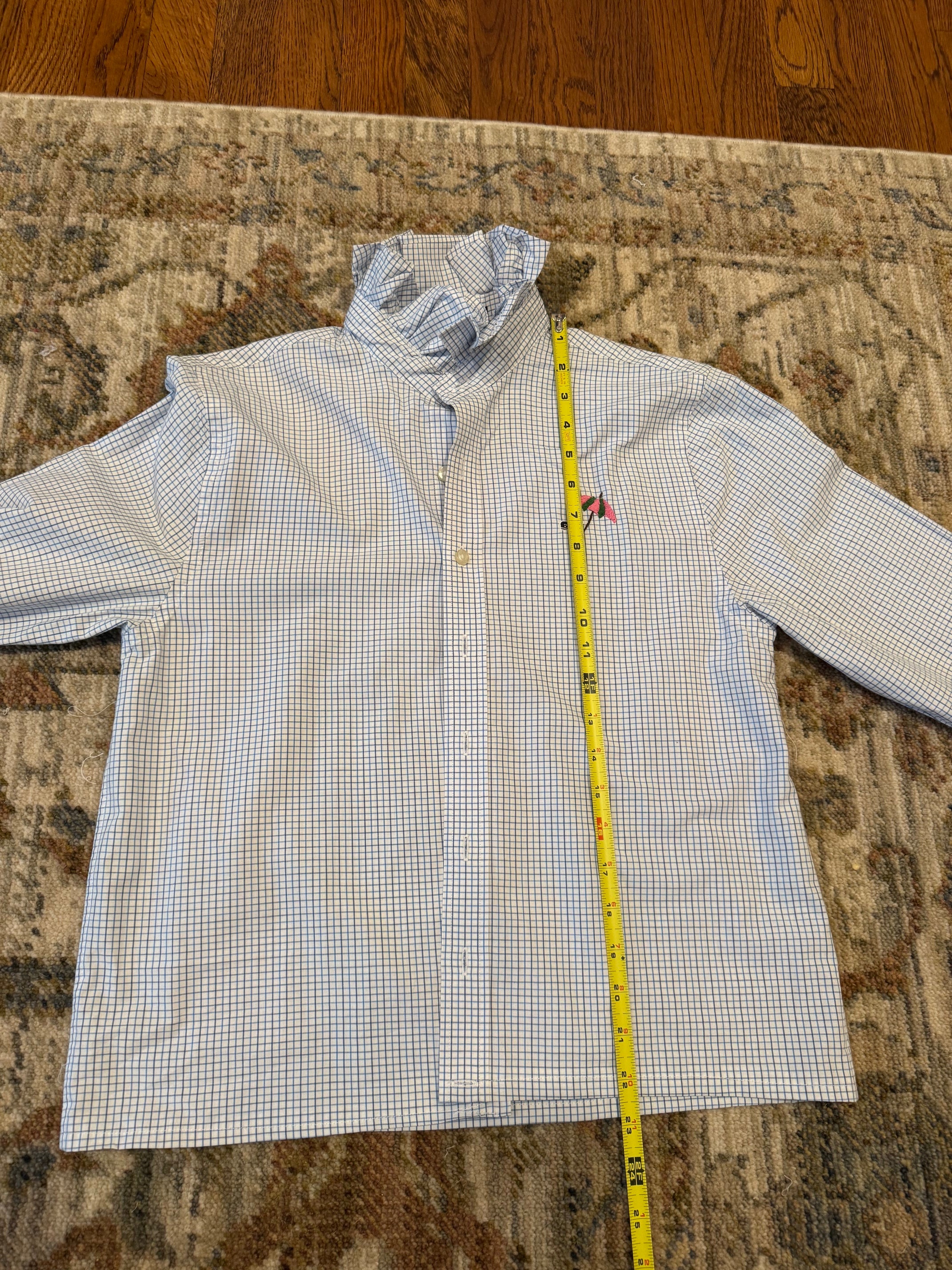 A long-sleeve button-up shirt laid flat on a patterned rug with a yellow tape measure standing vertically next to it for size reference.