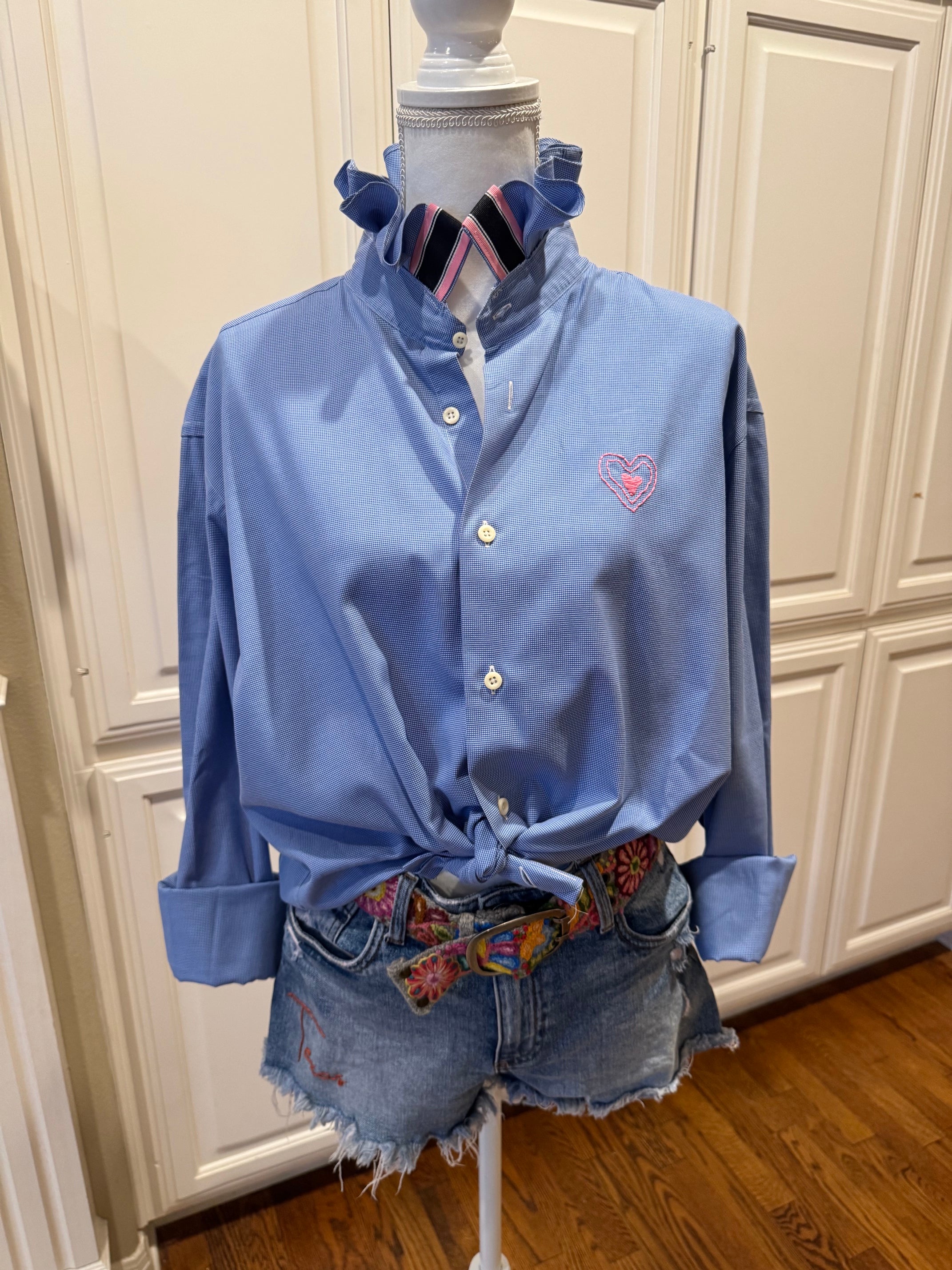 A light blue long-sleeve button-up shirt with a pink embroidered heart on the chest, styled on a mannequin with a striped shirt collar, distressed denim shorts, and a colorful patterned belt in front of white paneled cabinets.