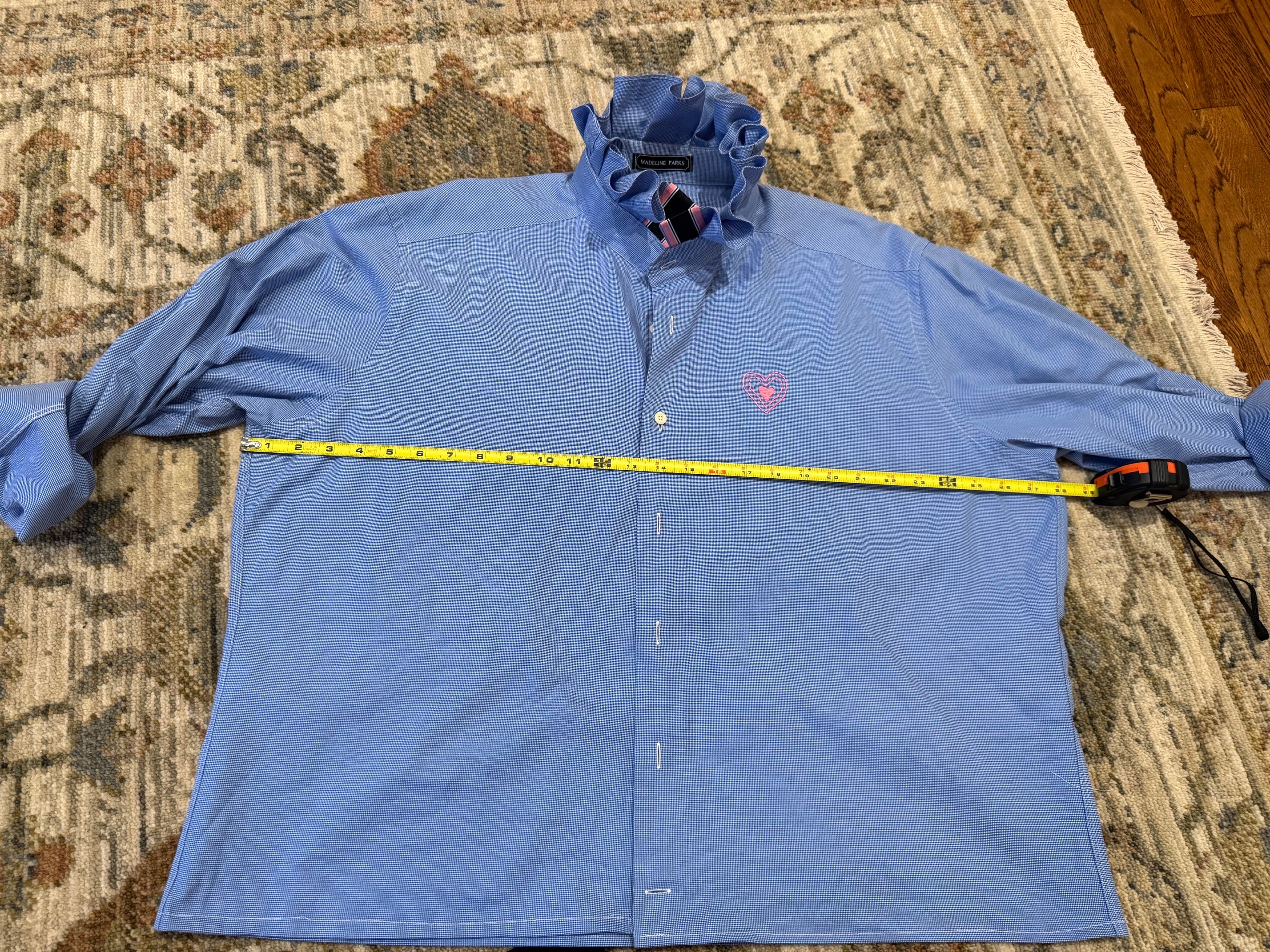 A light blue long-sleeve button-up shirt laid flat on a patterned rug, with a yellow tape measure stretched horizontally across the chest to show the width. The shirt has a small pink heart logo on the left chest.