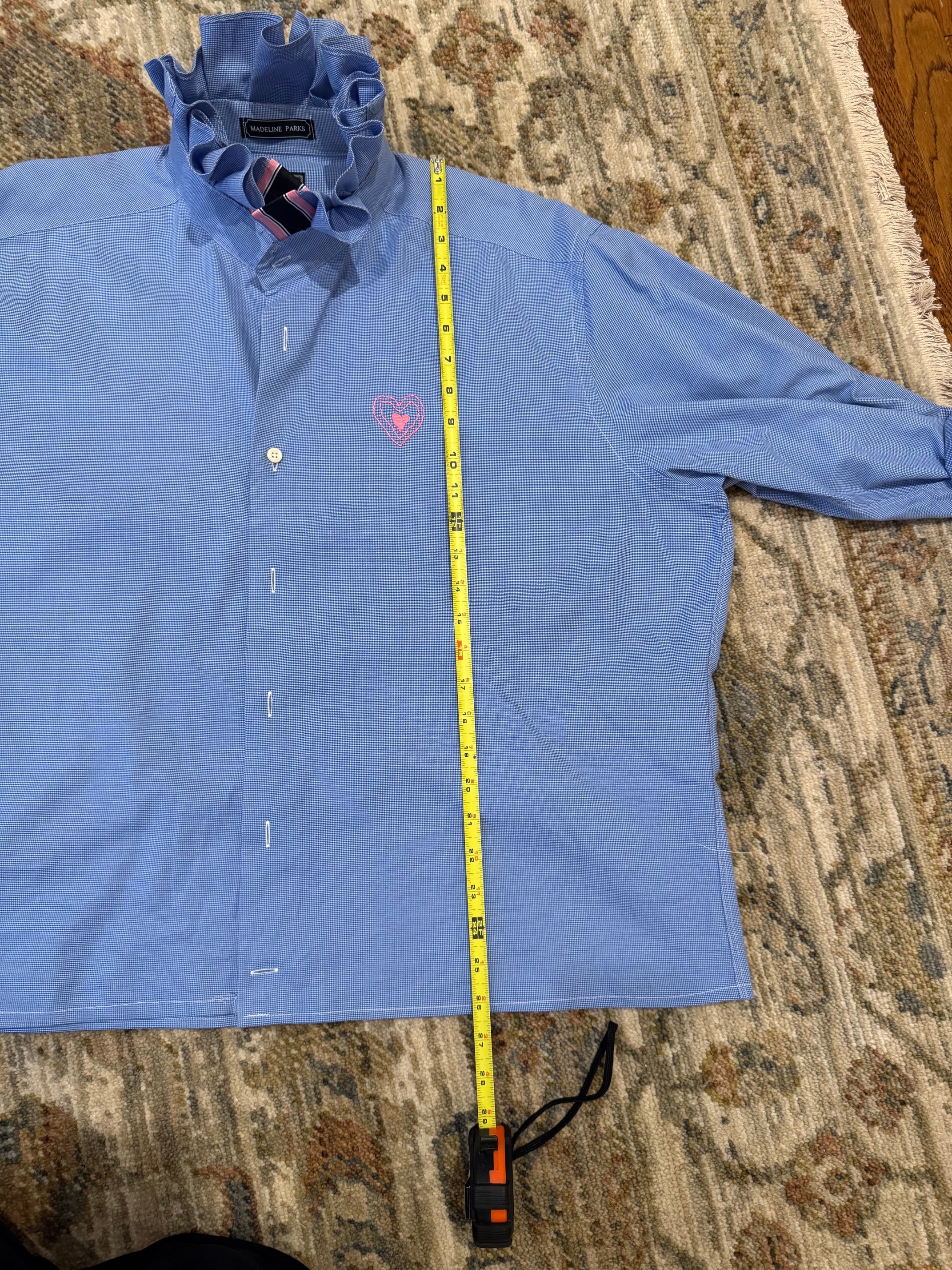 A light blue long-sleeve button-up shirt laid flat on a patterned rug, with a yellow tape measure placed vertically along the right side to show the shirt's length.