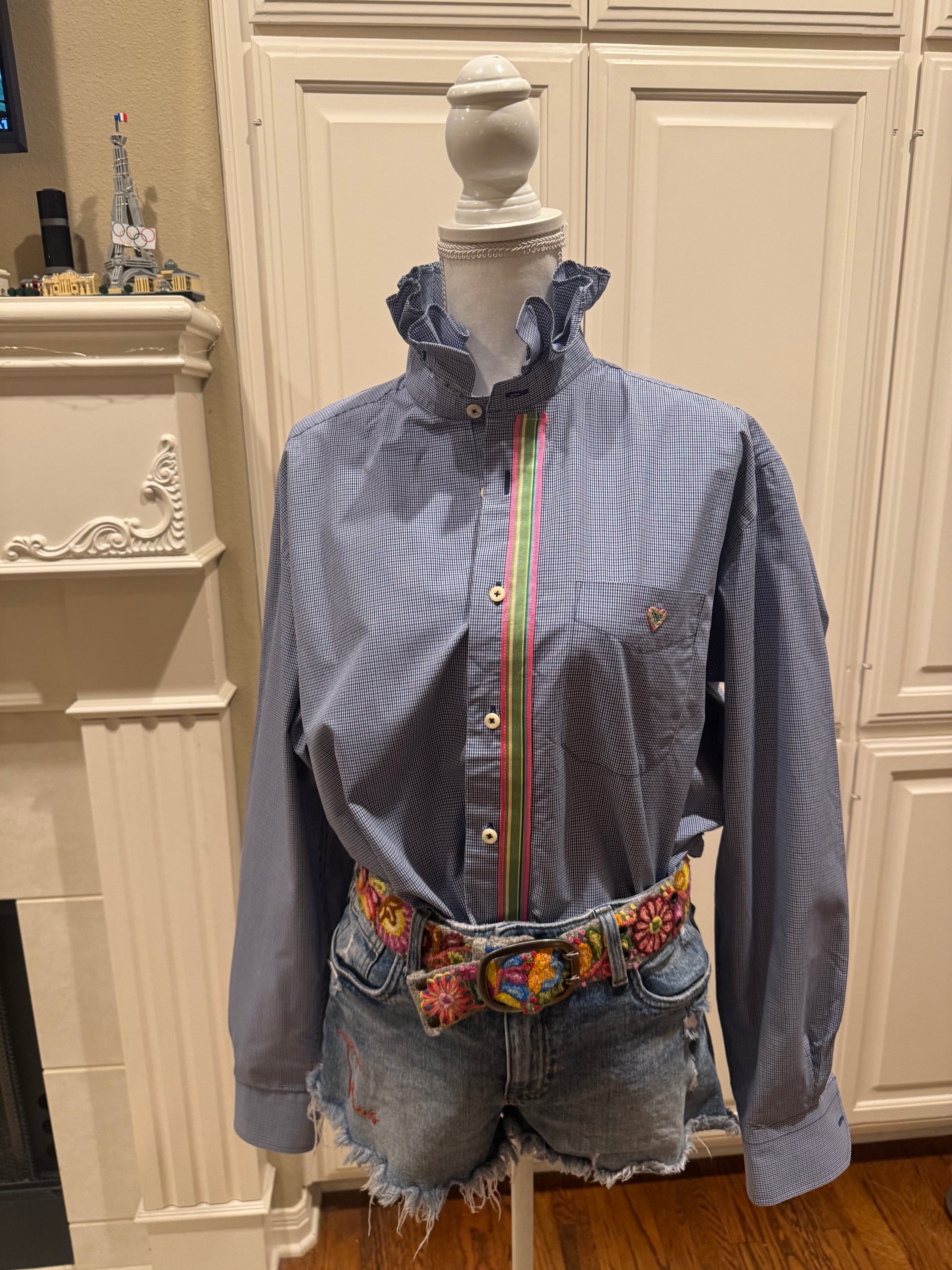 A blue long-sleeve button-up shirt with a ruffled high collar and a colorful vertical stripe detail down the front, styled on a mannequin with denim shorts and a patterned belt in front of a cream-colored cabinet background.