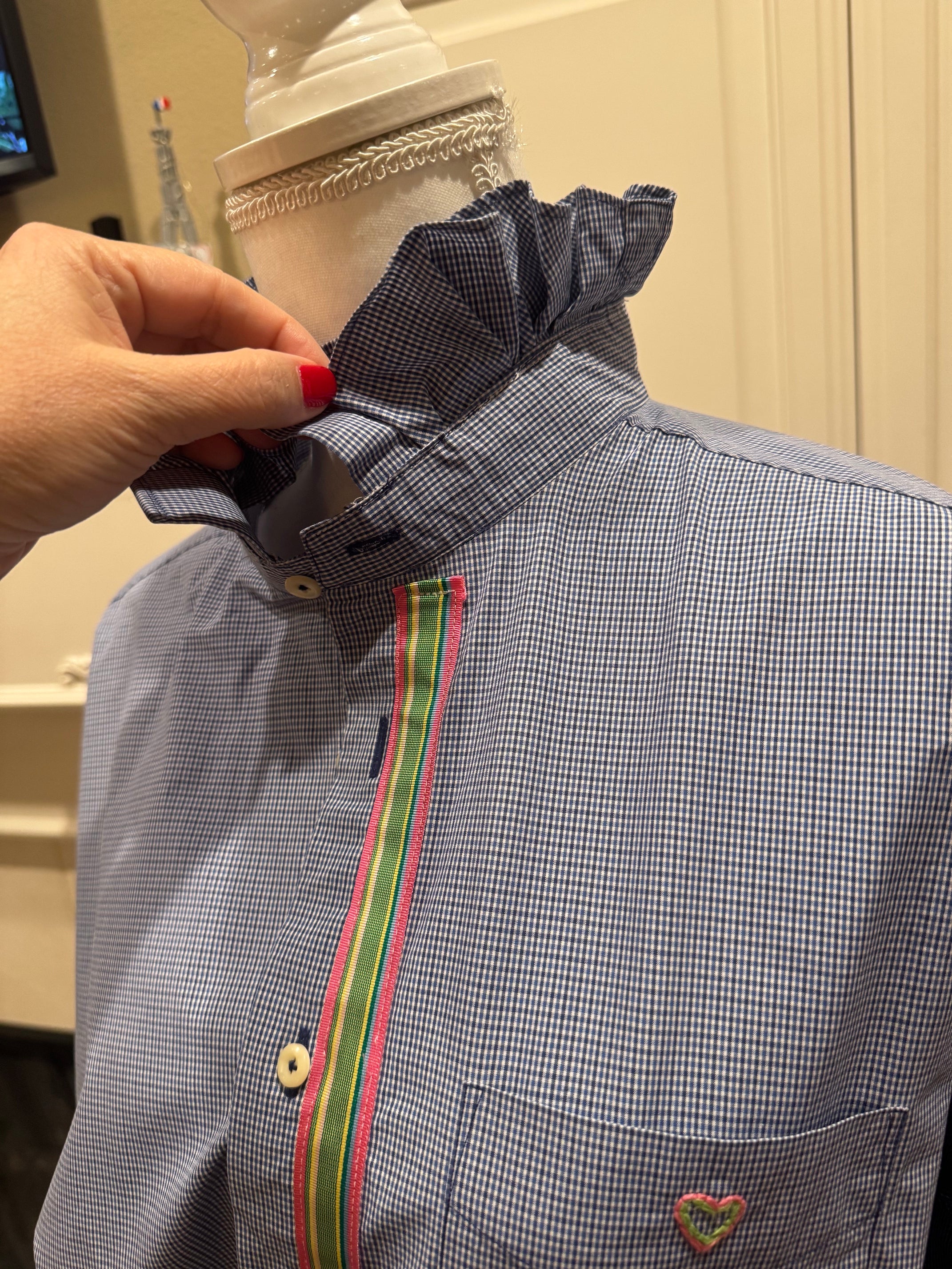 A close-up of a blue and white micro-check shirt on a mannequin, featuring a ruffled collar being adjusted by a hand with red nail polish, a vertical green and pink stripe down the front, and a small embroidered heart patch on the chest pocket.