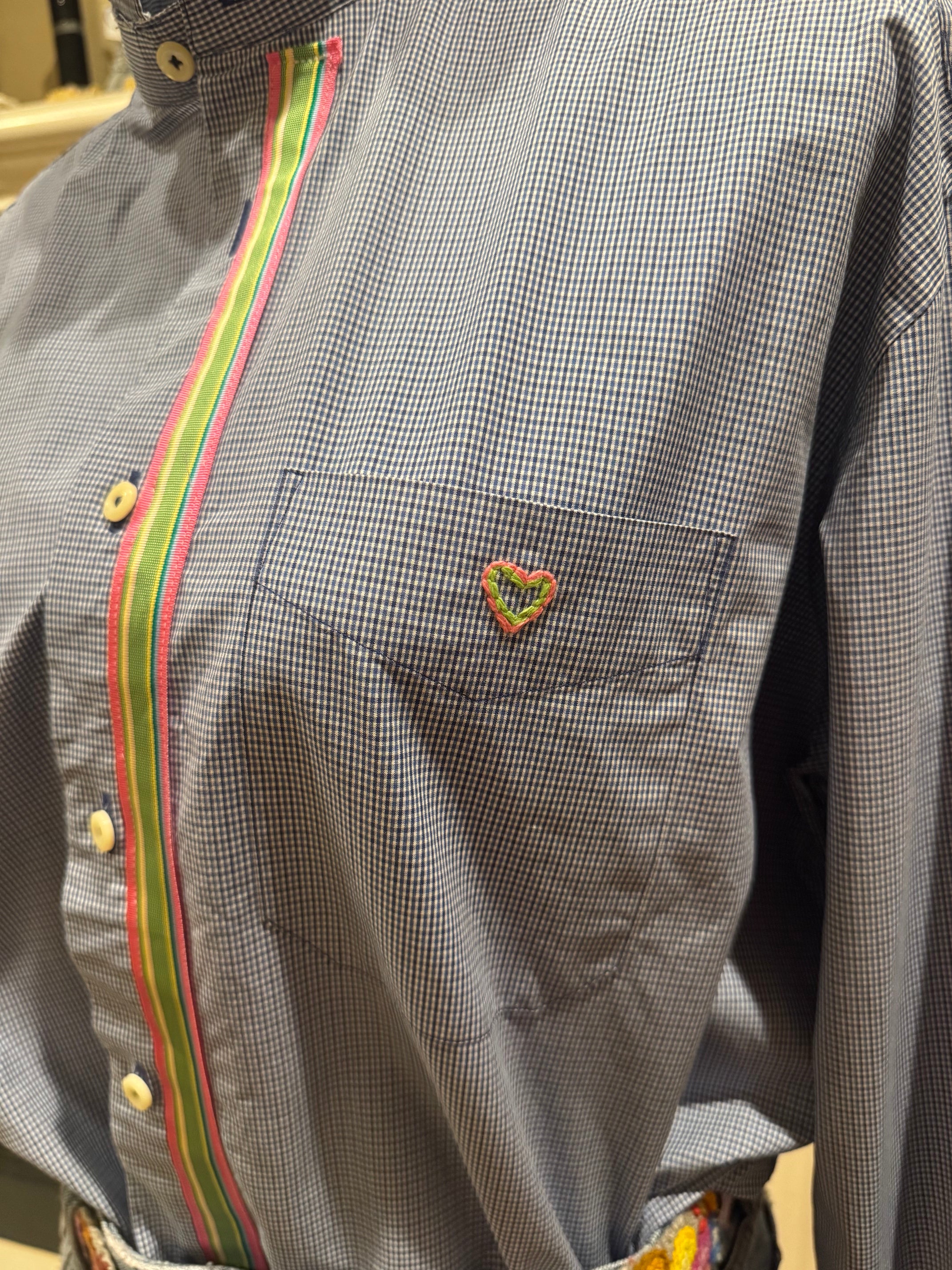 A close-up of a long-sleeve button-up shirt with a fine gray and white micro-check pattern, featuring a vertical stripe accent in green, yellow, and red along the placket and collar. A small embroidered heart patch is visible on the chest pocket.
