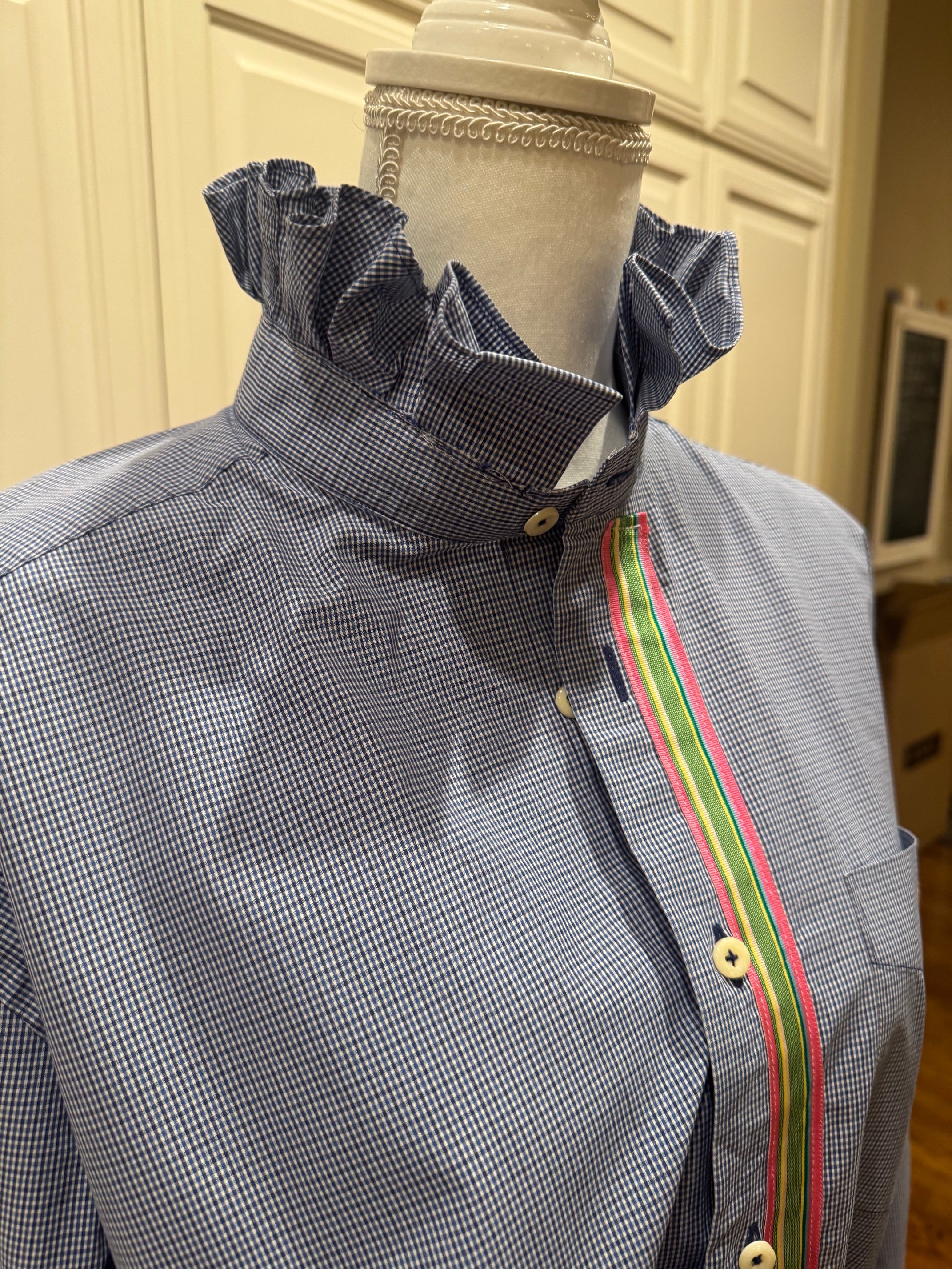A close-up of a blue and white micro-check patterned shirt with a ruffled high collar, displayed on a mannequin. The shirt features a vertical decorative stripe on the front with green, yellow, and red borders, and is shown against a background of cream-colored wooden cabinets.