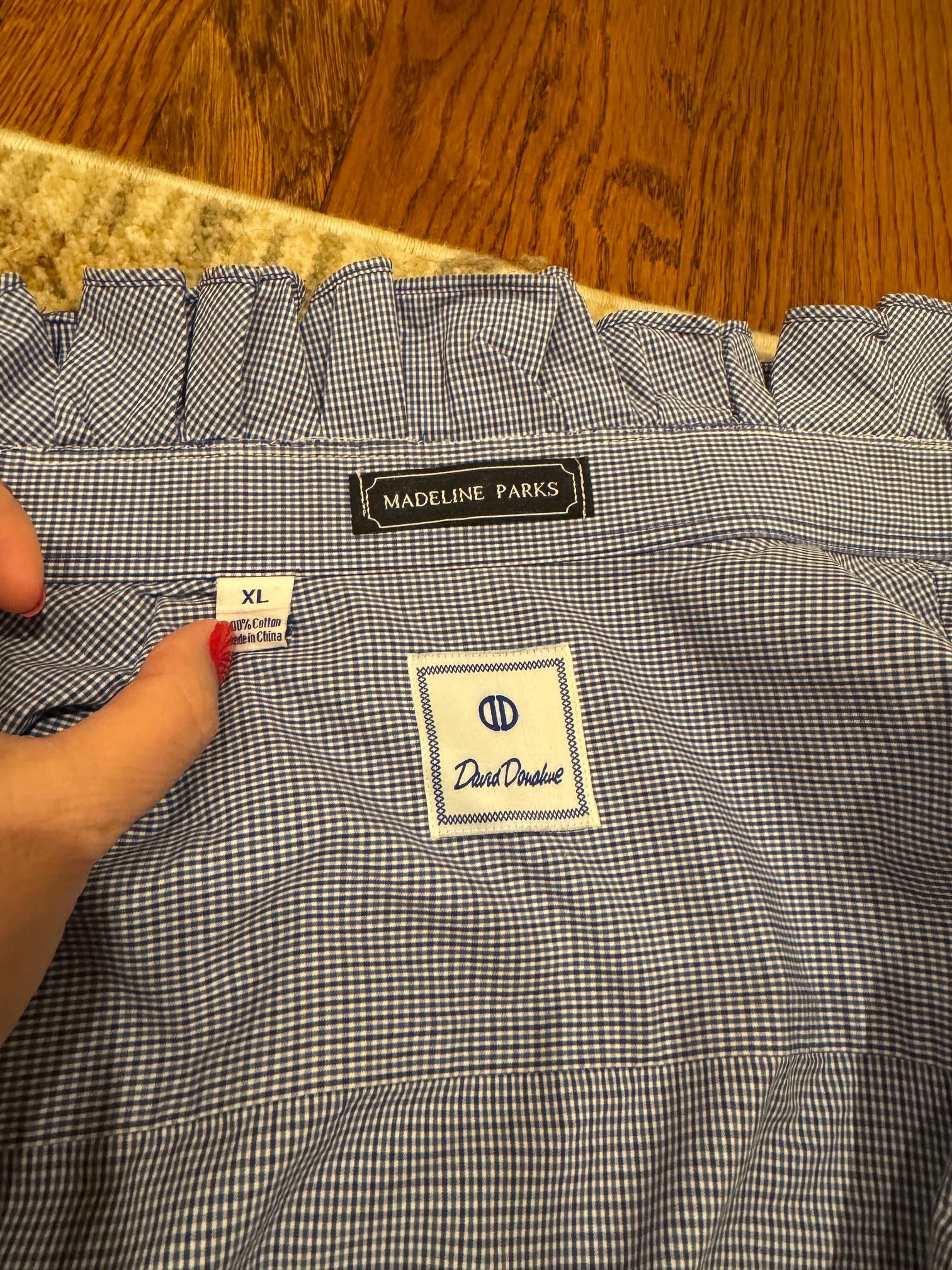 Close-up of the inside back of a blue and white gingham blouse, showing a ruffled neckline, a Madeline Parks brand label, a David Dickie brand patch, and a hand holding the fabric.