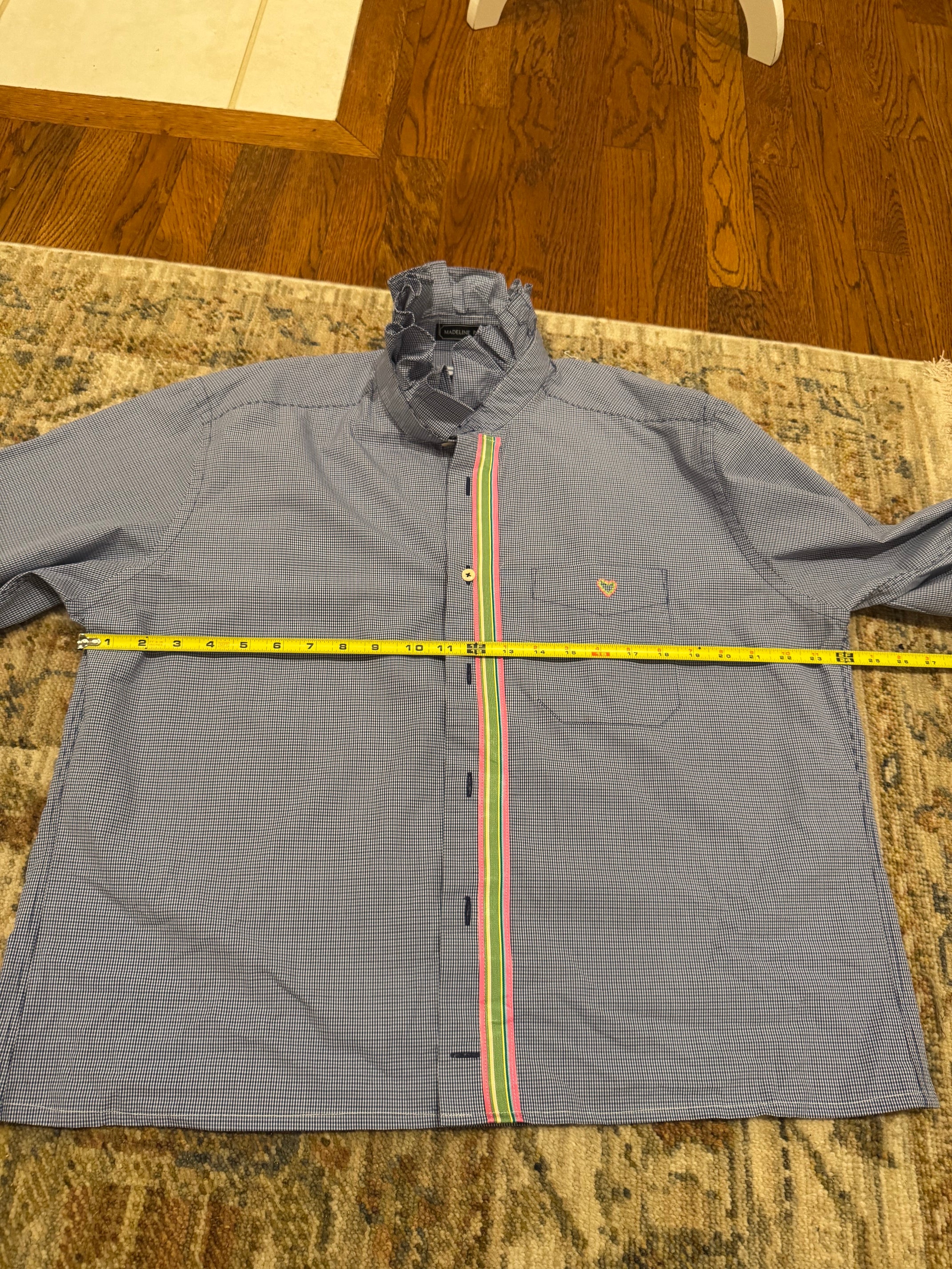 A gray long-sleeve button-up shirt laid flat on a patterned rug with a yellow tape measure spanning the chest. The shirt has a small heart logo on the left chest and a vertical multicolor stripe detail down the front.