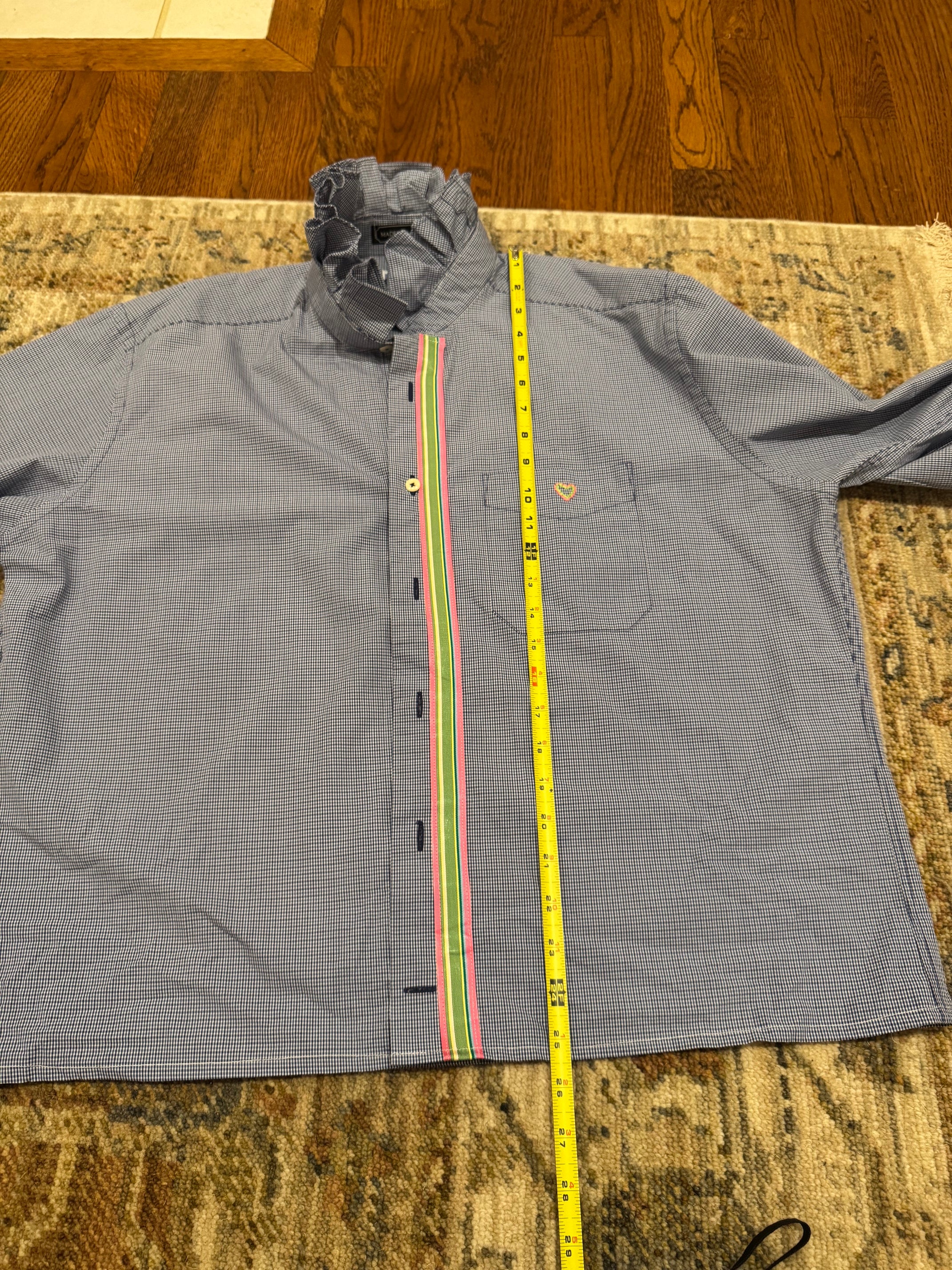 A gray long-sleeve button-up shirt laid flat on a patterned rug, with a yellow tape measure placed vertically down the center front to show the garment's width.