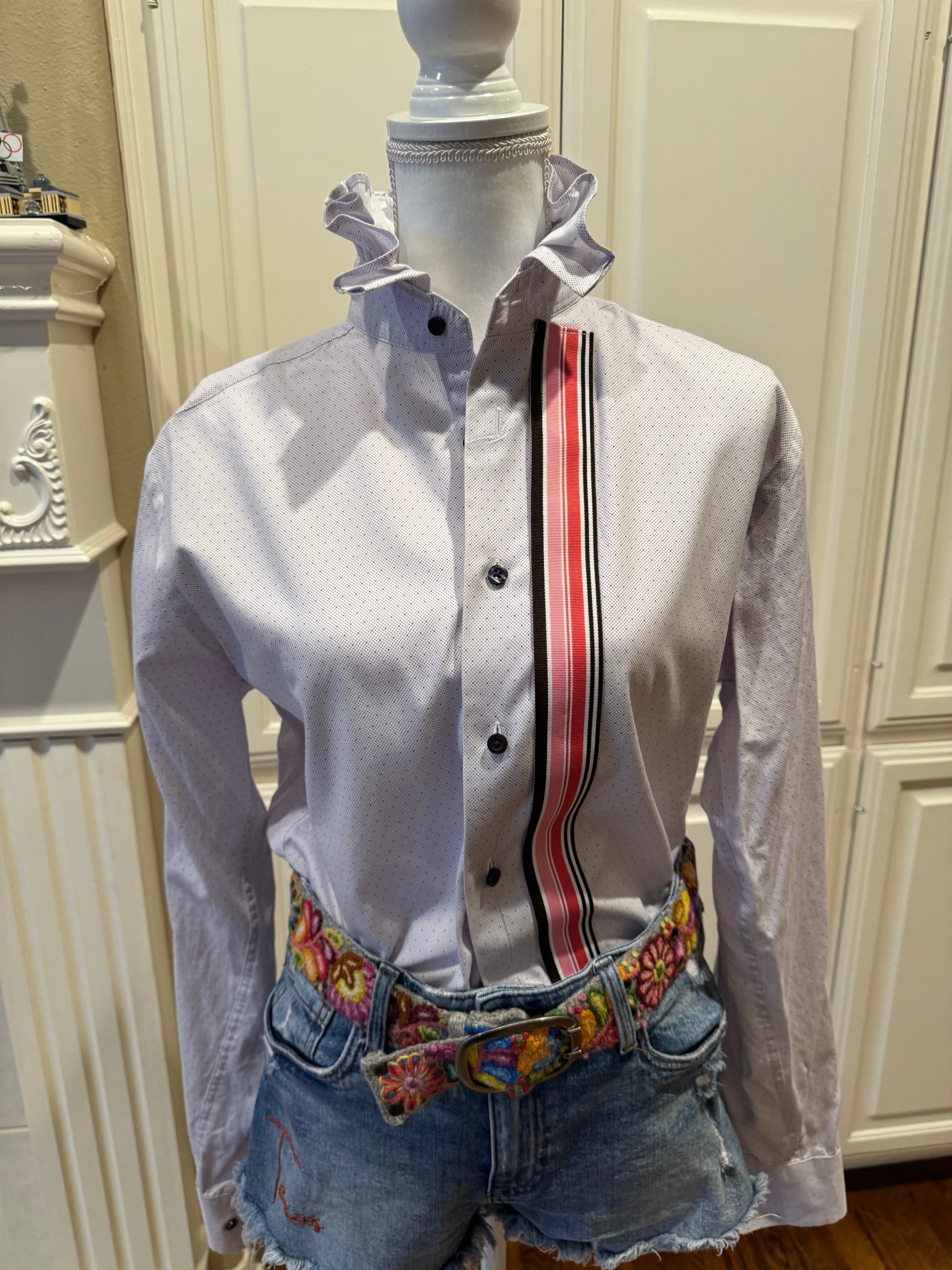 A white long-sleeve button-up shirt with a ruffled collar and a vertical red, black, and white stripe detail on the chest, styled on a mannequin with denim shorts and a colorful embroidered belt.