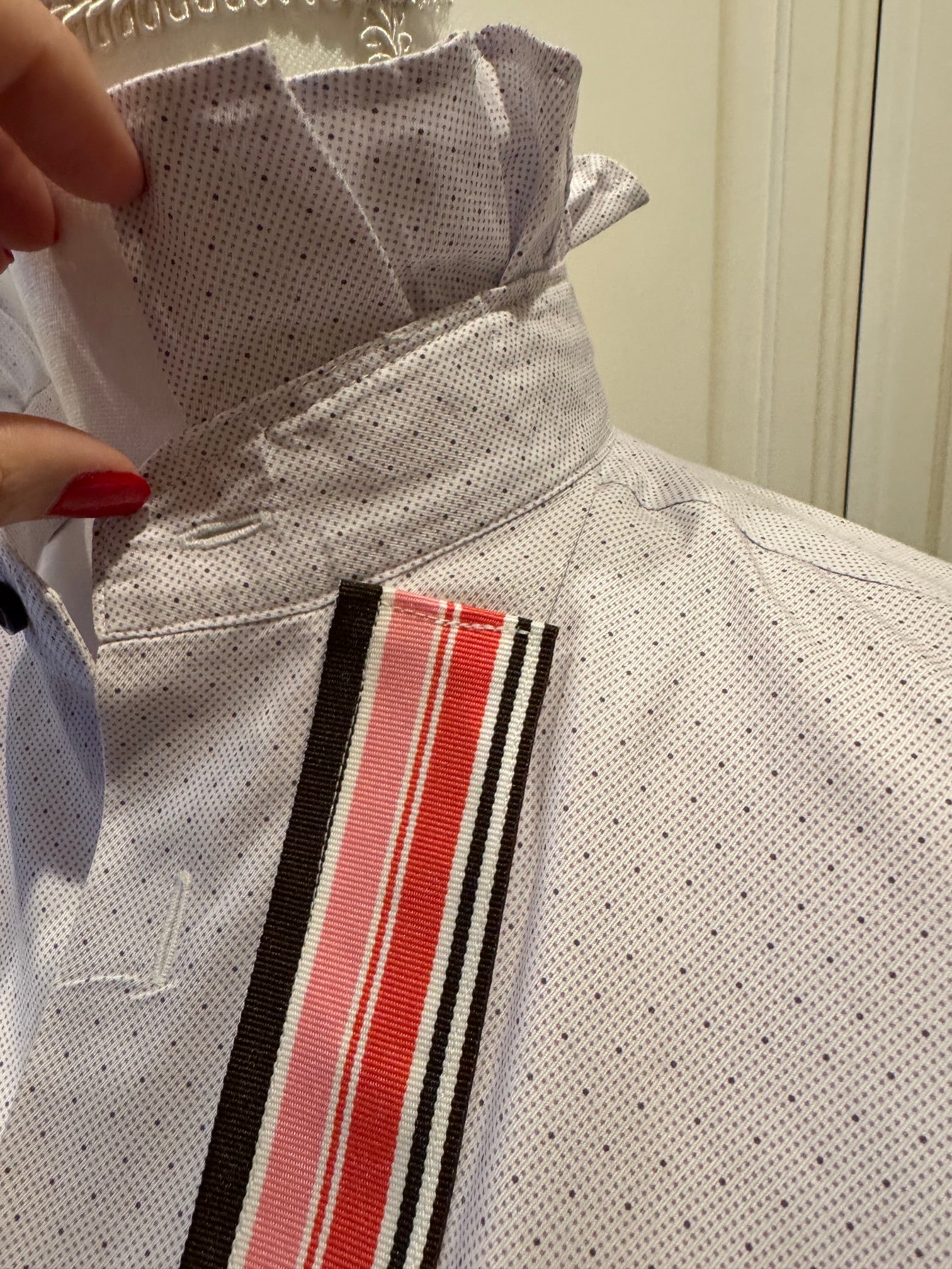 A close-up of a white short-sleeve dress shirt with a fine micro-dot pattern, featuring a high pleated collar and a vertical striped ribbon detail in black, white, and red.