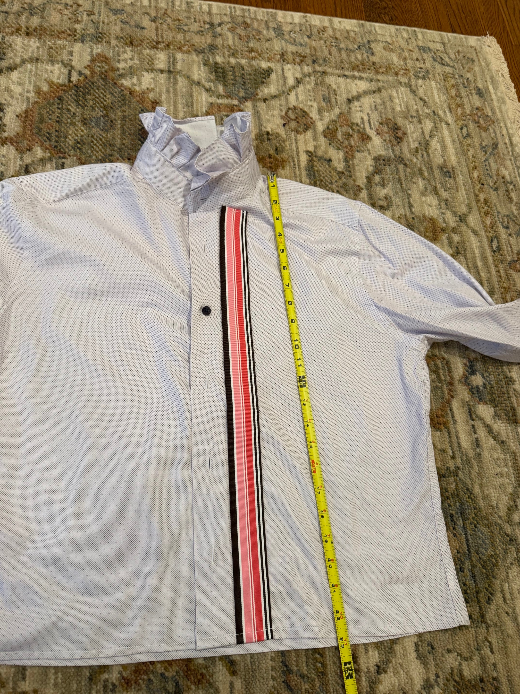 A white long-sleeve dress shirt with a vertical striped placket in black, red, and white, laid flat on a patterned rug with a yellow measuring tape running vertically to show the shirt's length.