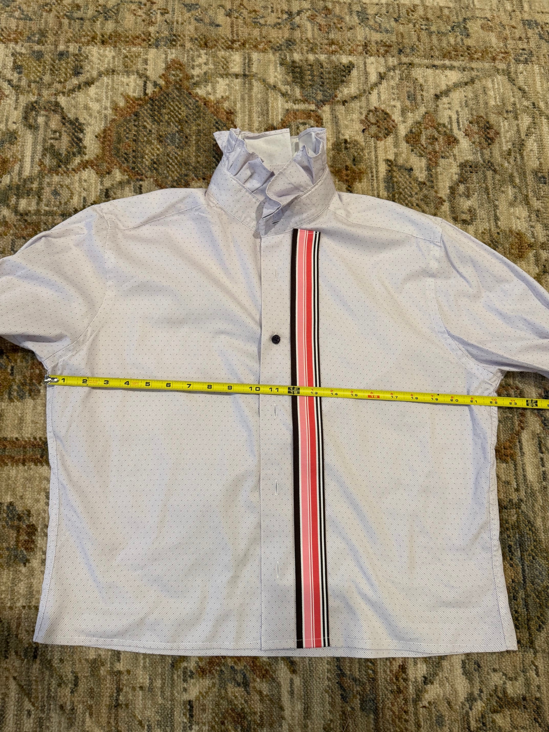A white long-sleeve button-up shirt with a high collar and a vertical stripe detail in red, pink, and black down the front, laid flat on a patterned rug with a yellow measuring tape across the chest.