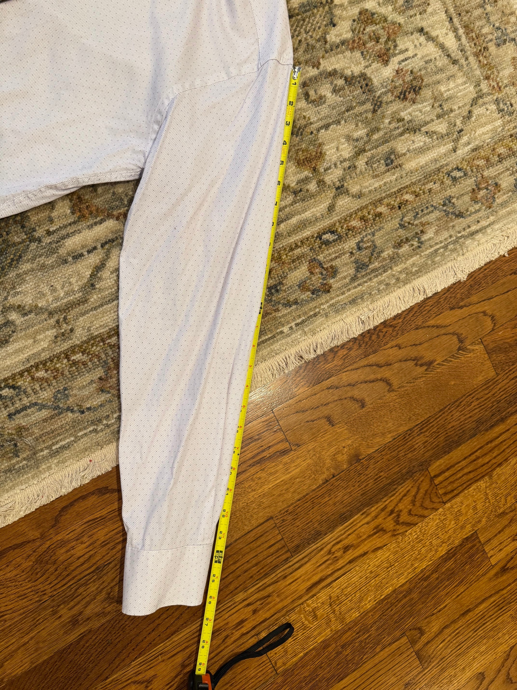 A light-colored dress shirt laid flat on a patterned rug over a hardwood floor, with a yellow tape measure running vertically along the sleeve to show the length.