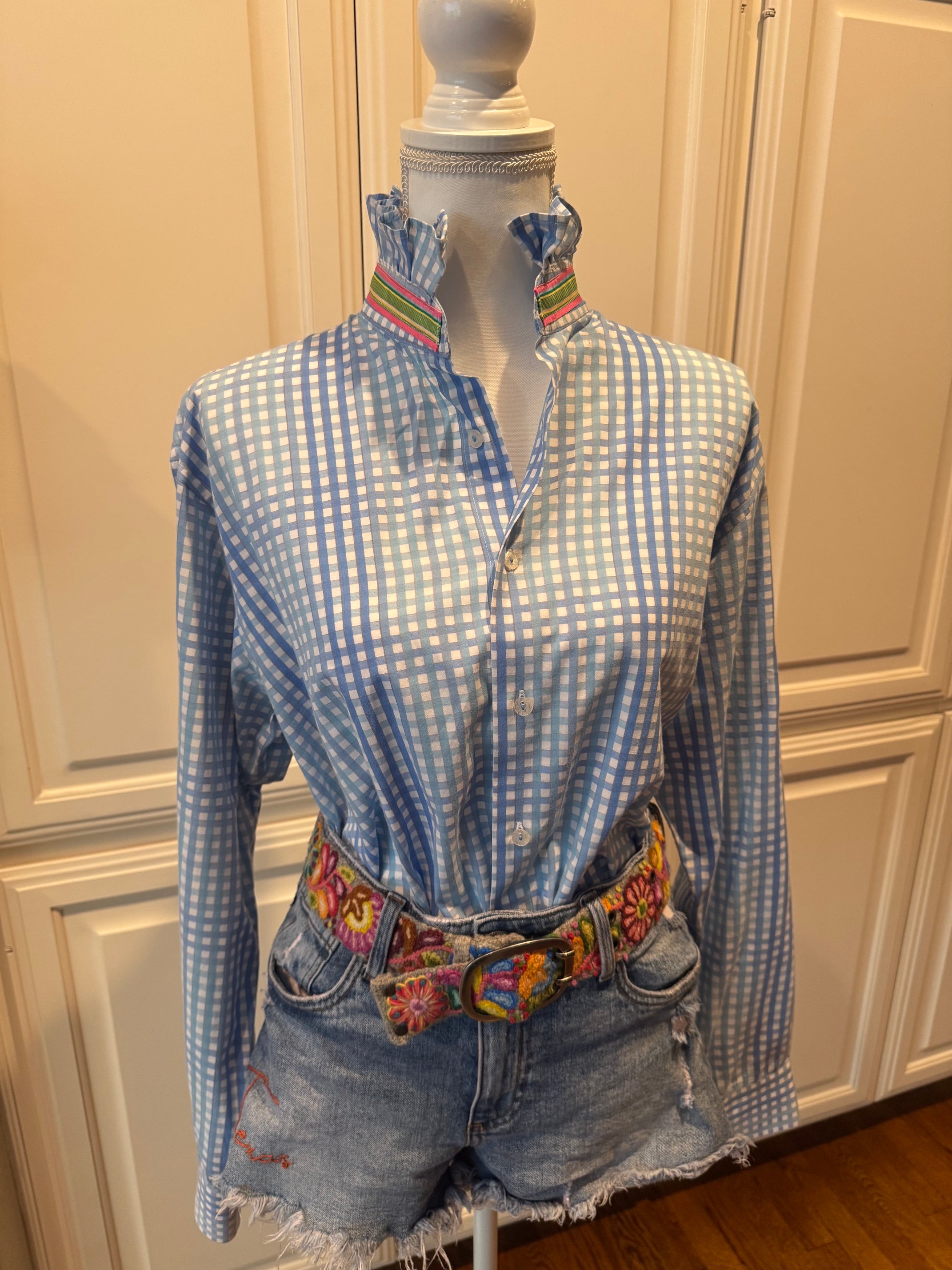 A blue and white gingham long-sleeve shirt with a colorful embroidered collar, styled on a mannequin with denim shorts and a matching embroidered belt in front of cream-colored paneled cabinets.