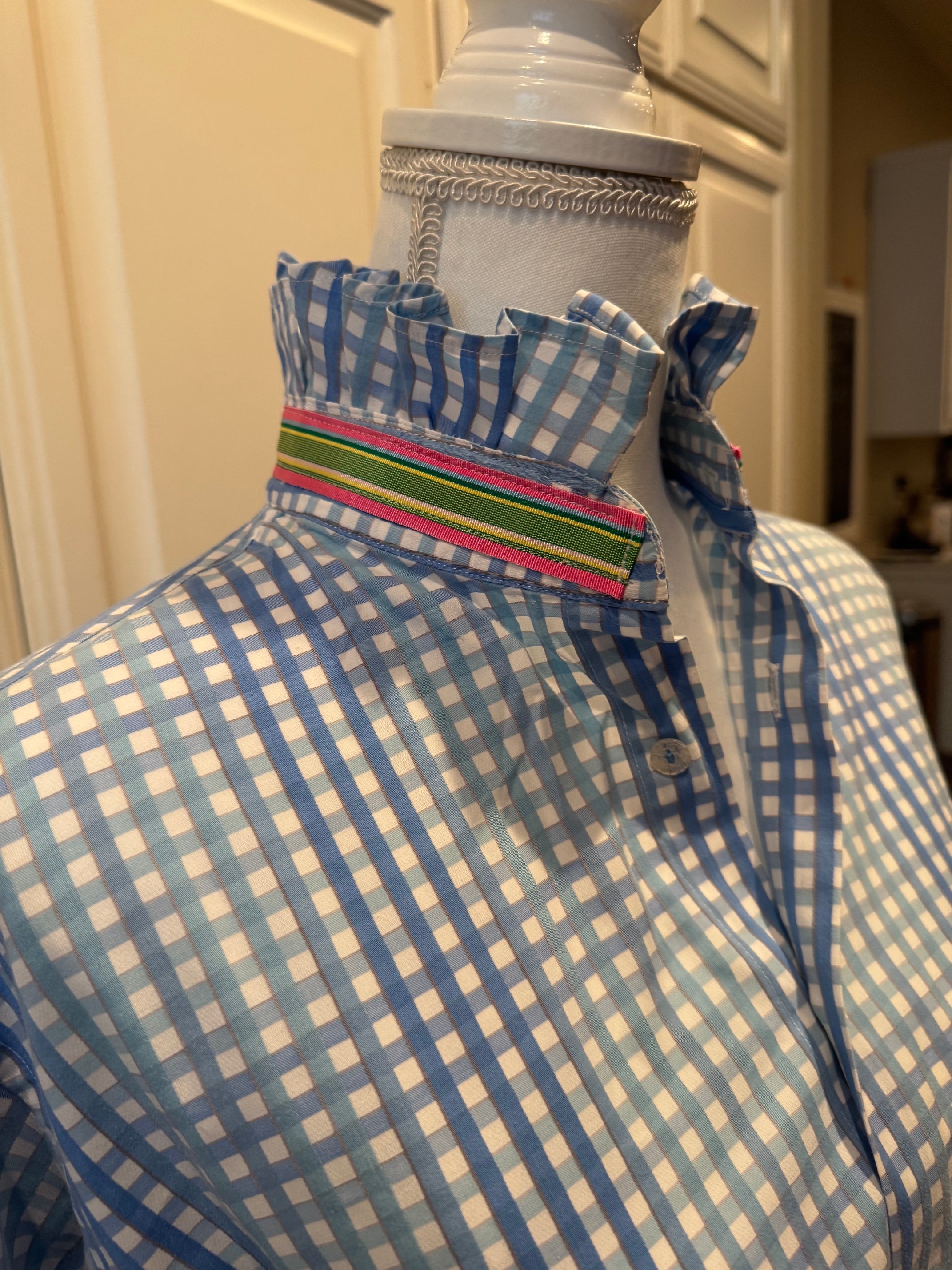 A close-up of a blue and white gingham button-down shirt with a ruffled collar and a colorful striped inner placket, displayed on a mannequin torso.