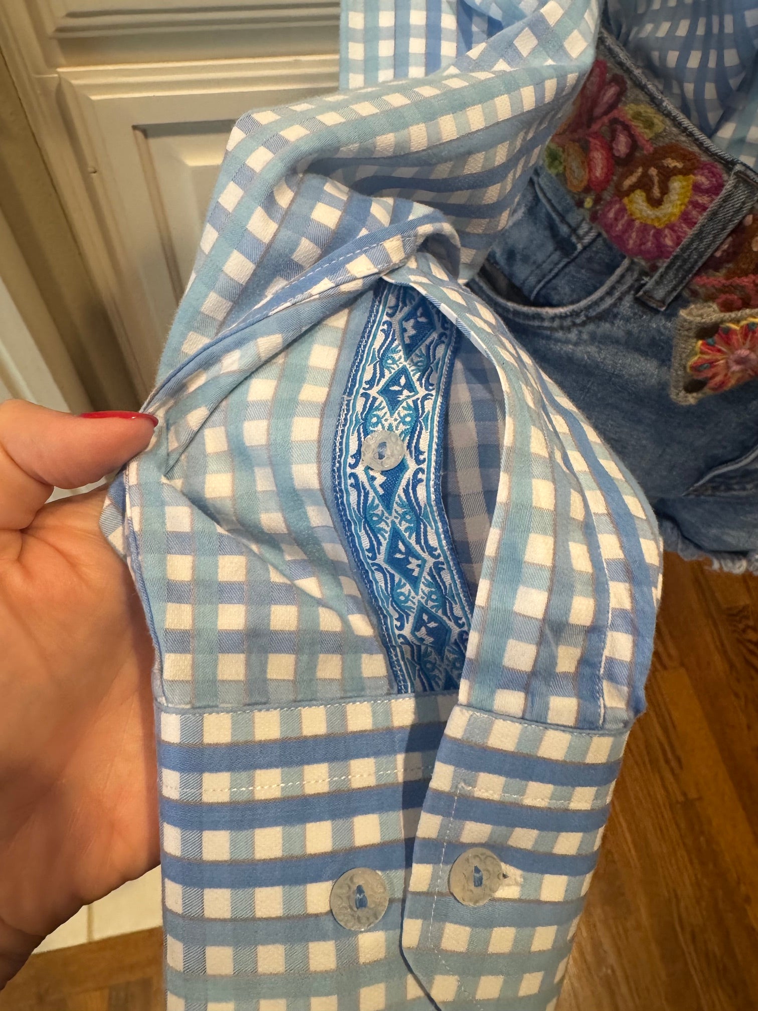 A close-up of a light blue and white gingham button-down shirt being held by a hand, showing its collar with a blue paisley embroidered inner band and front buttons.
