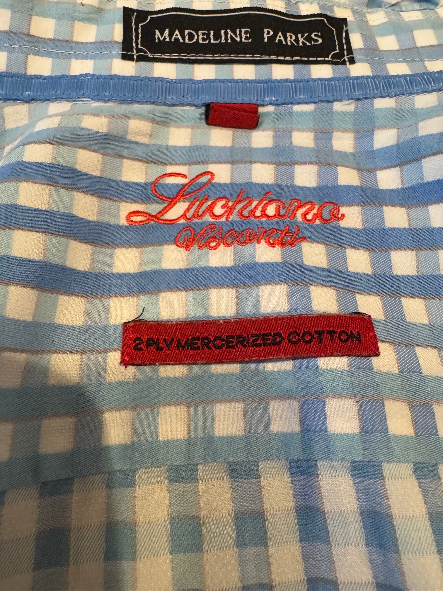 Close-up of a blue and white gingham checkered garment with red embroidered text reading 'Lucciano Visconti' and a red fabric label reading '2PLY MERCERIZED COTTON'. A black neck label reads 'MADELINE PARKS'.
