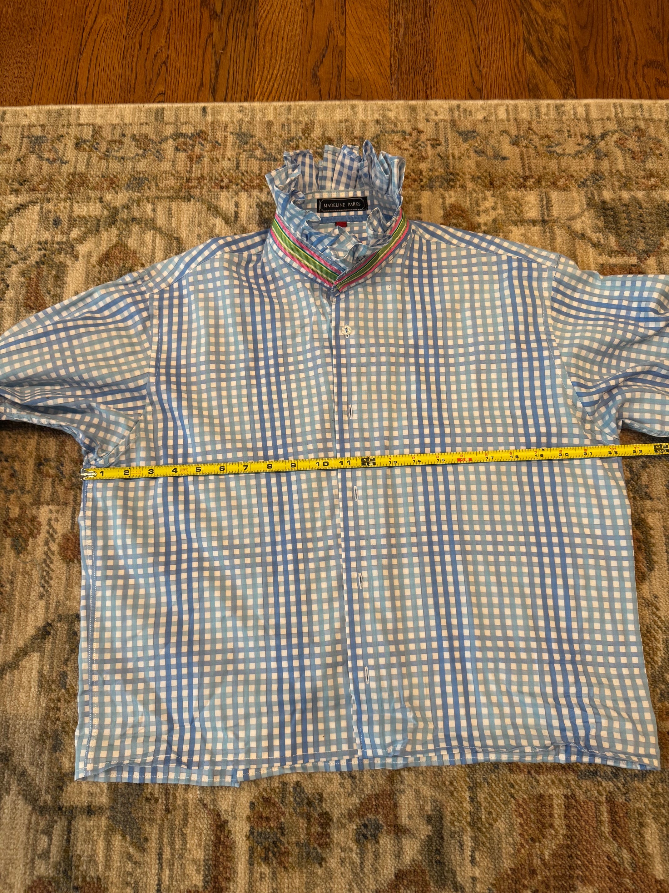A light blue and white gingham long-sleeve button-up shirt laid flat on a patterned rug, with a yellow tape measure stretched across the chest to show the width. The shirt has a striped collar and a visible neck label that reads 'BONSAI'.