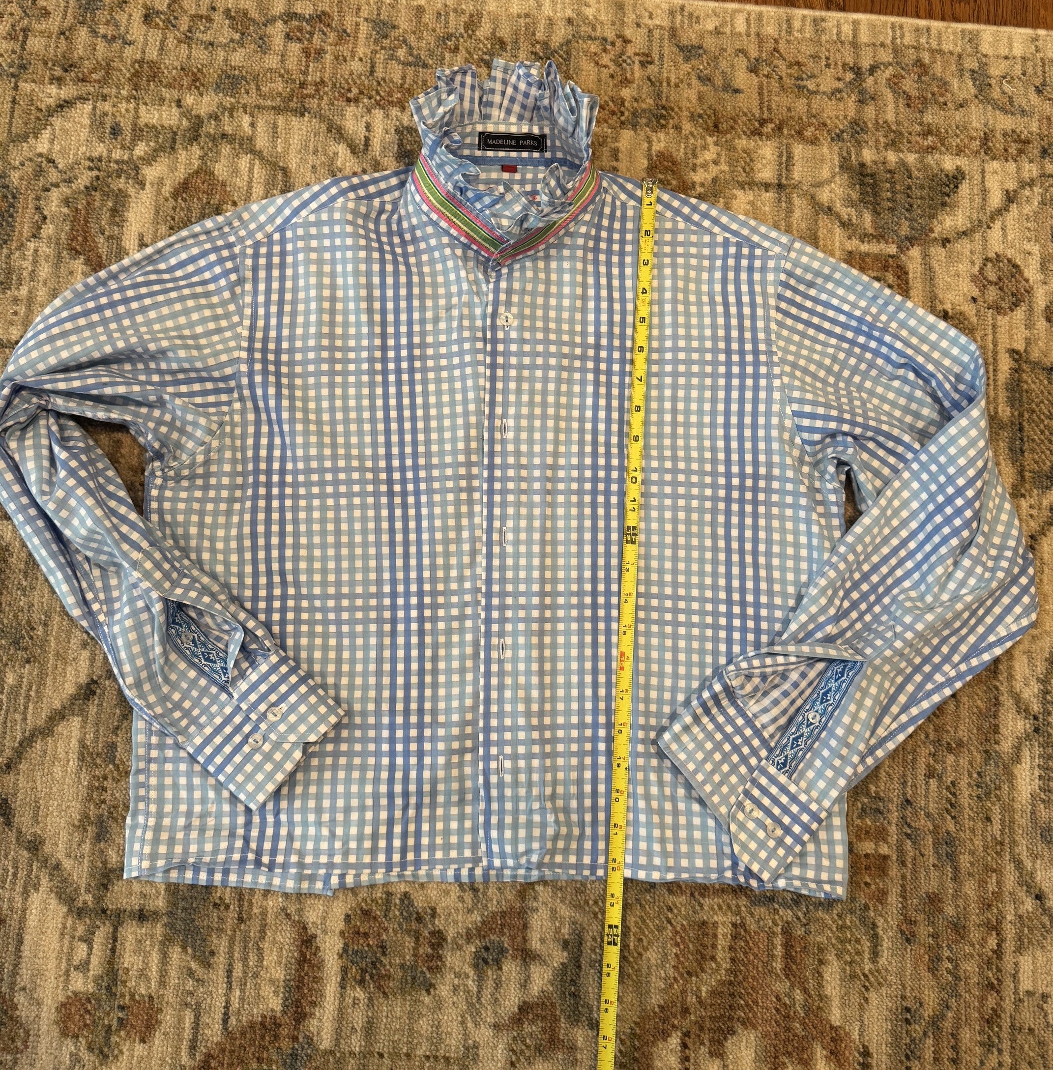 A men's long-sleeve button-up dress shirt with a light blue and white gingham pattern, laid flat on a patterned area rug with a yellow tape measure standing vertically to indicate size.