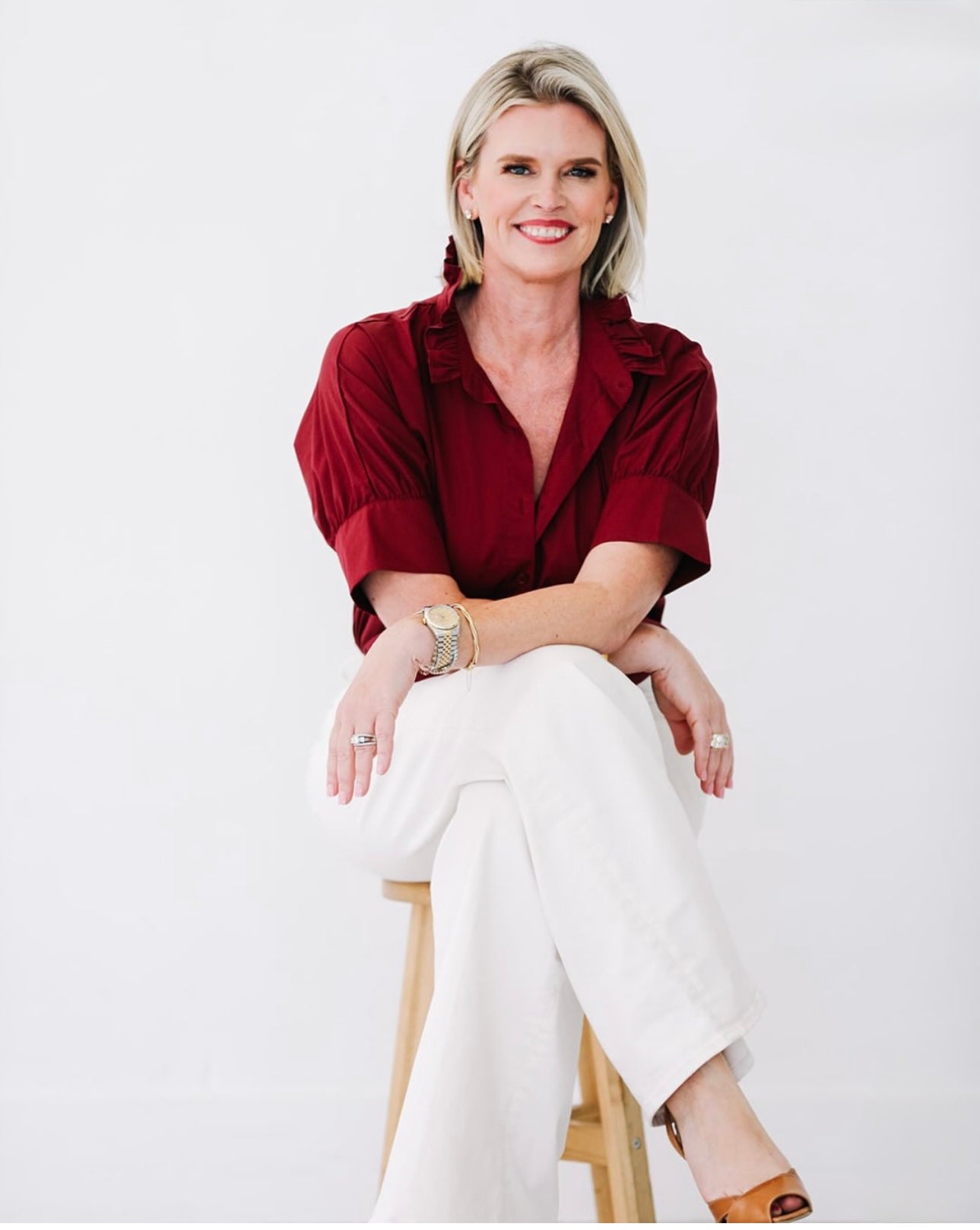 Woman wearing Heidi Houston Ruffle Shirt in Maroon, seated, showcasing the blouse's subtle ruffle collar detail, paired with white pants and tan heels.