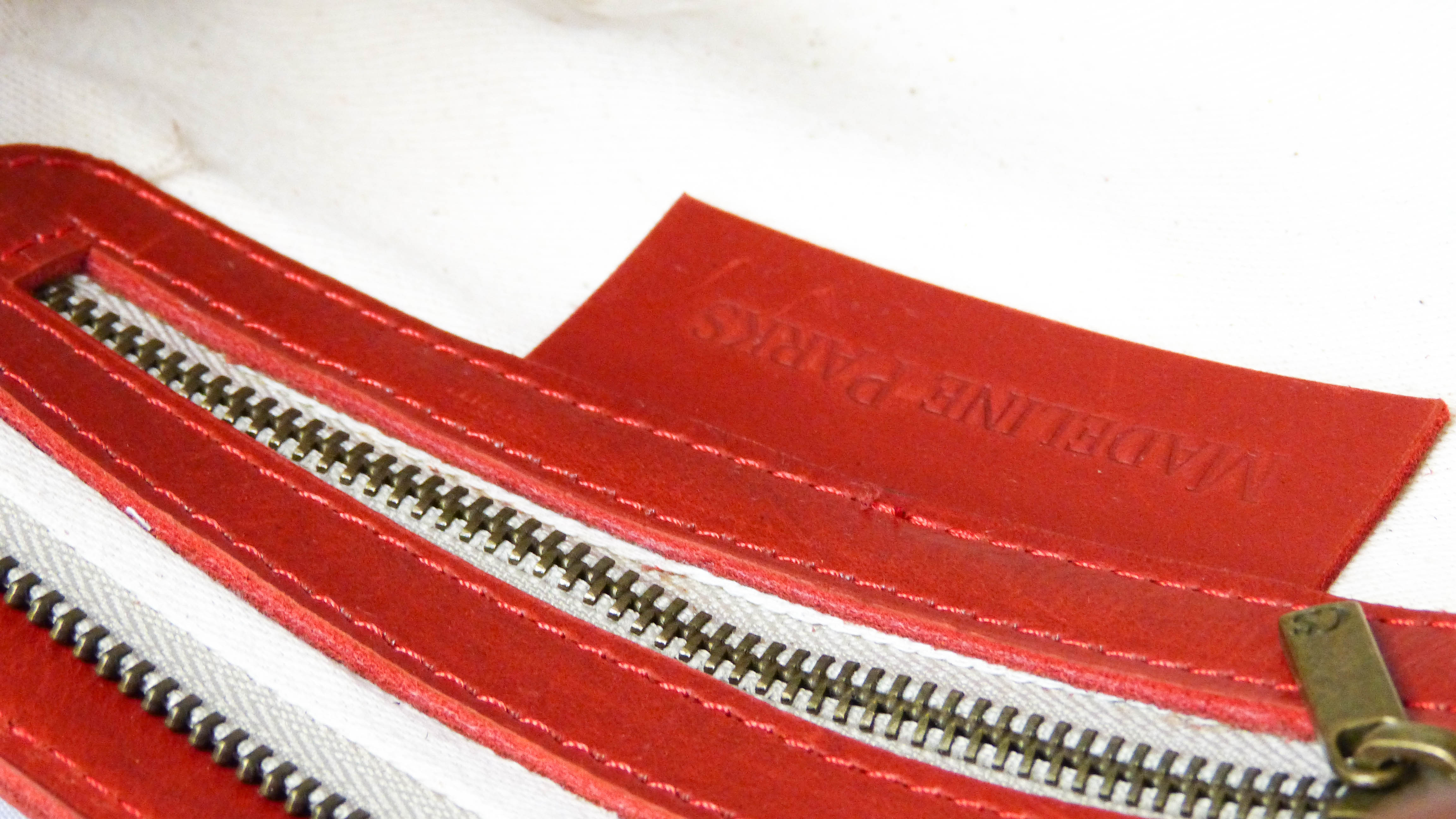 Close-up of the Jessica Leather Purse in Cherry, showcasing a white fabric with a red leather label, a zipper, and intricate stitching. Handmade by Ethiopian artisans for Madeline Parks Designs.