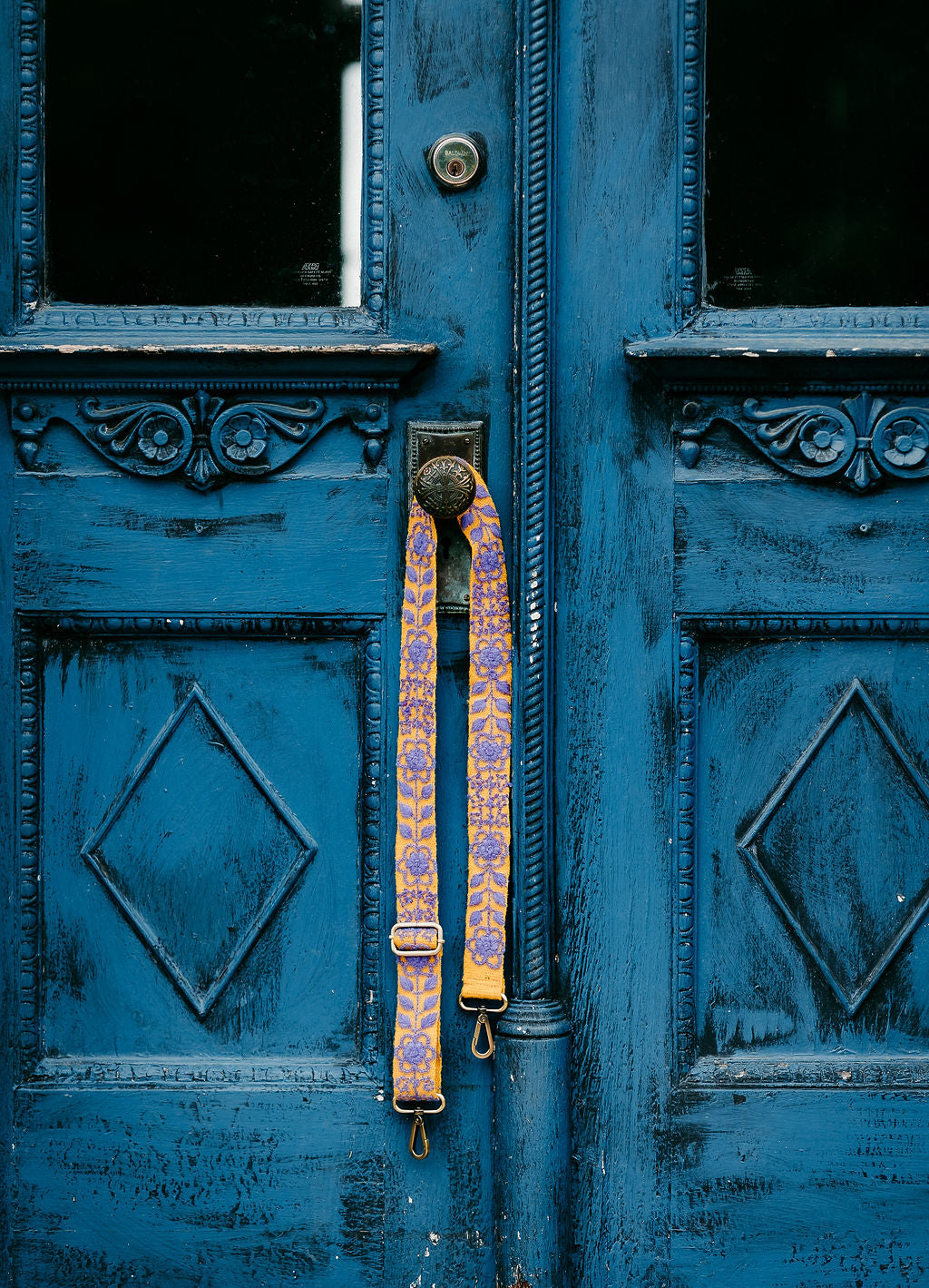 Arden Adjustable Strap: Hand-woven wool with floral and leaves design Shown hanging on a doorknob of an antique blue door.