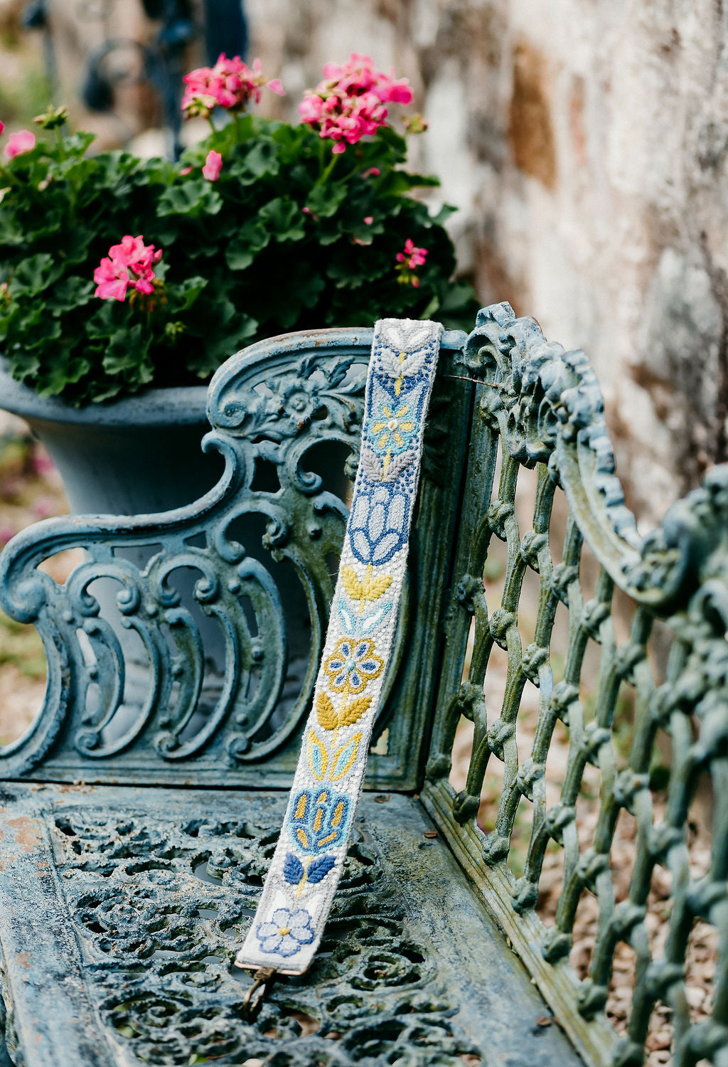 A purse strap with a floral and leaves design in yellow, purple, and grey displayed on a bench