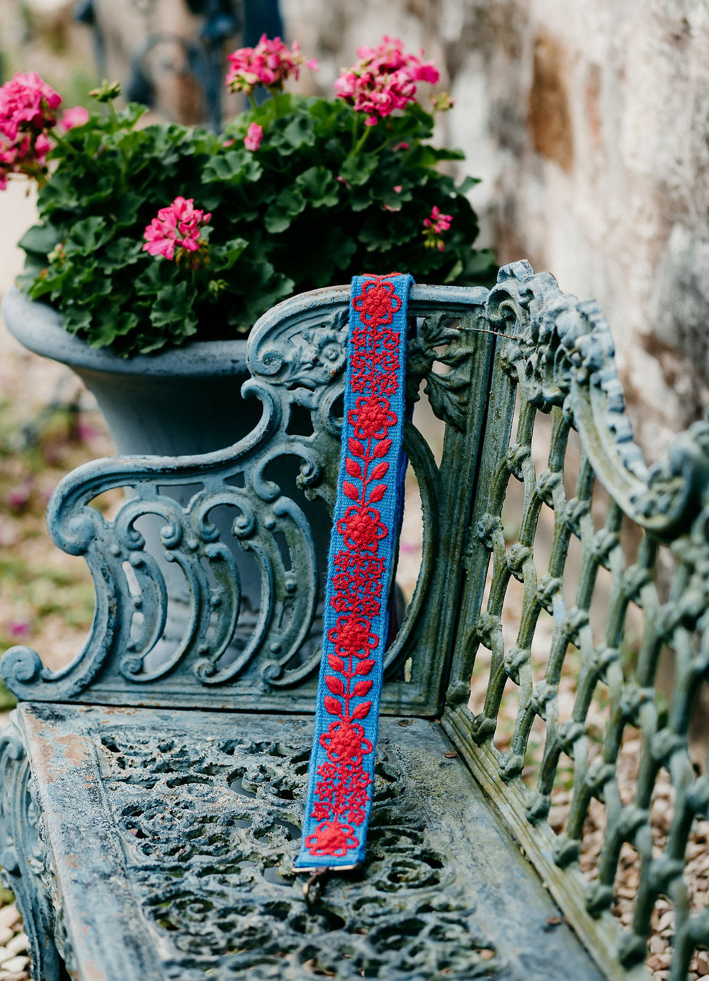 Red and blue purse strap adorned with red floral and leaf embroidery displayed on a bench.