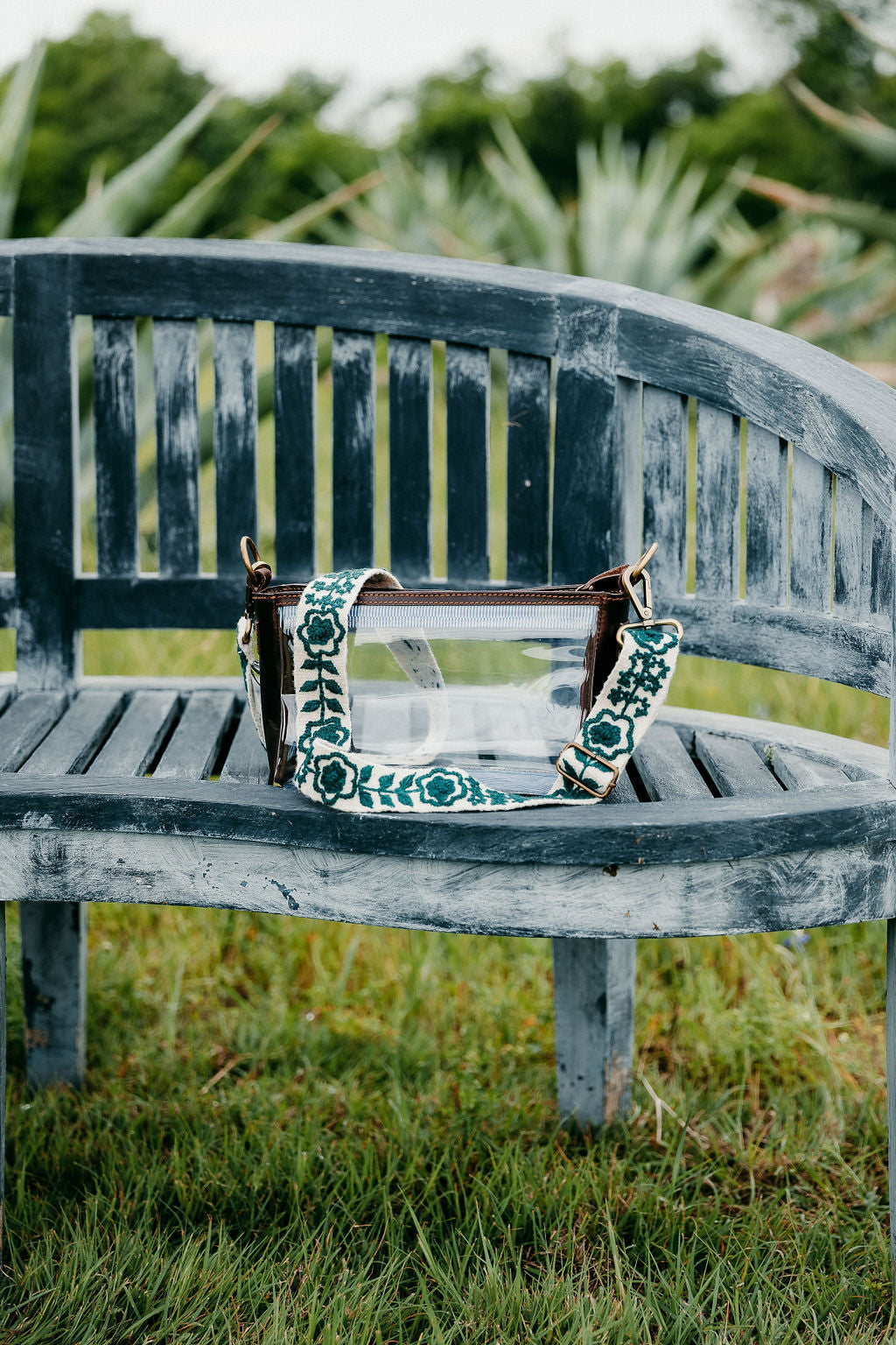 Jill Adjustable Strap, a clear bag with a green strap, on bench in an outdoor setting.
